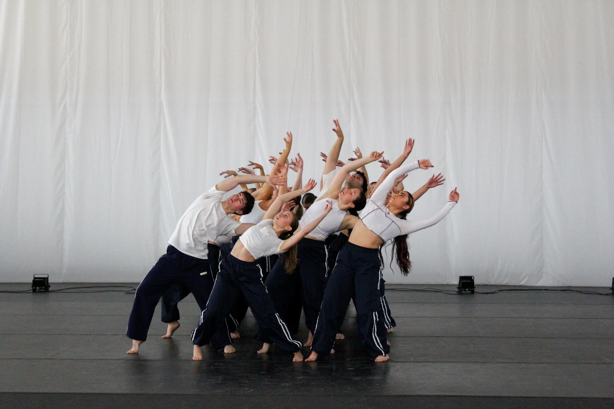 A group of dancers in athletic clothing performing a choreographed dance routine on stage. The dancers are standing in a line with some leaning back and reaching upward with their arms, against a plain, white backdrop.