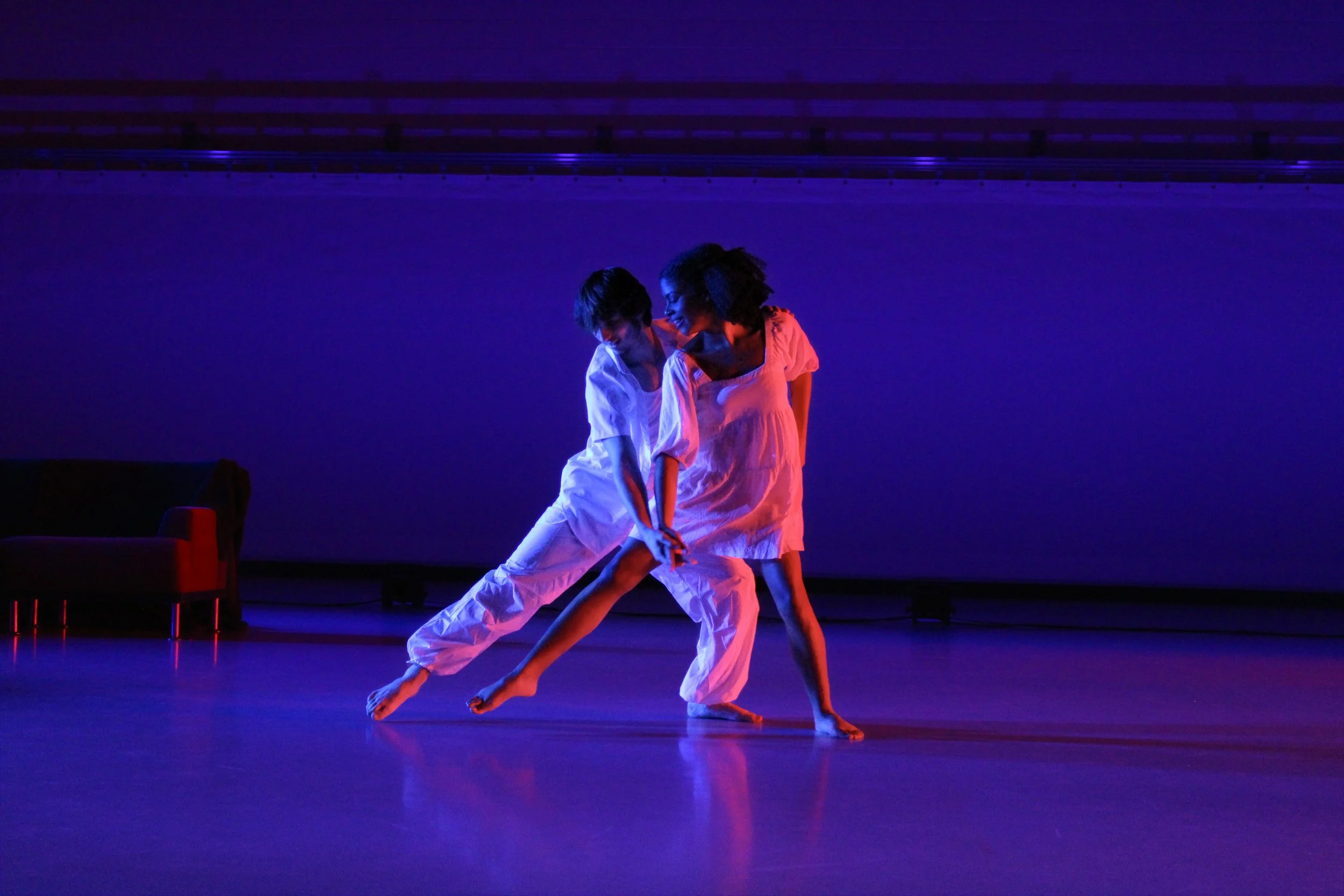 Two dancers with curly hair performing in a dark room illuminated by purple and blue lighting. They are dressed in white, one wearing a dress and the other in a shirt and pants, engaging in a dance movement with intertwined legs.