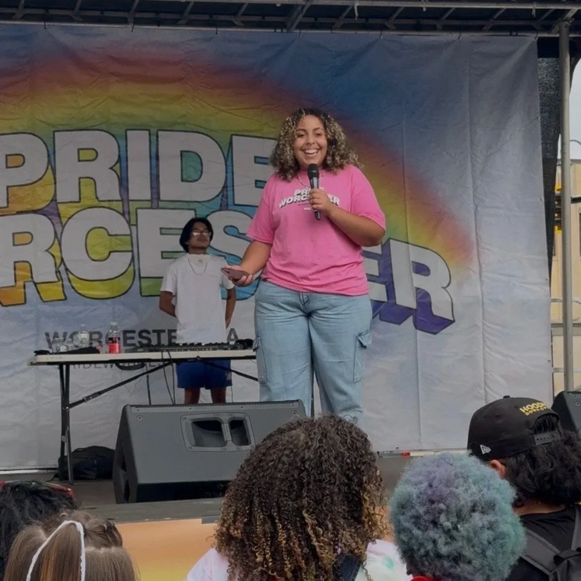 A woman smiling on stage at a pride event, holding a microphone, wearing a pink shirt with the words 'Pride Warrior,' with a rainbow-colored banner behind her reading 'Pride R' and 'WC' partially visible; a DJ or sound technician stands behind her.
