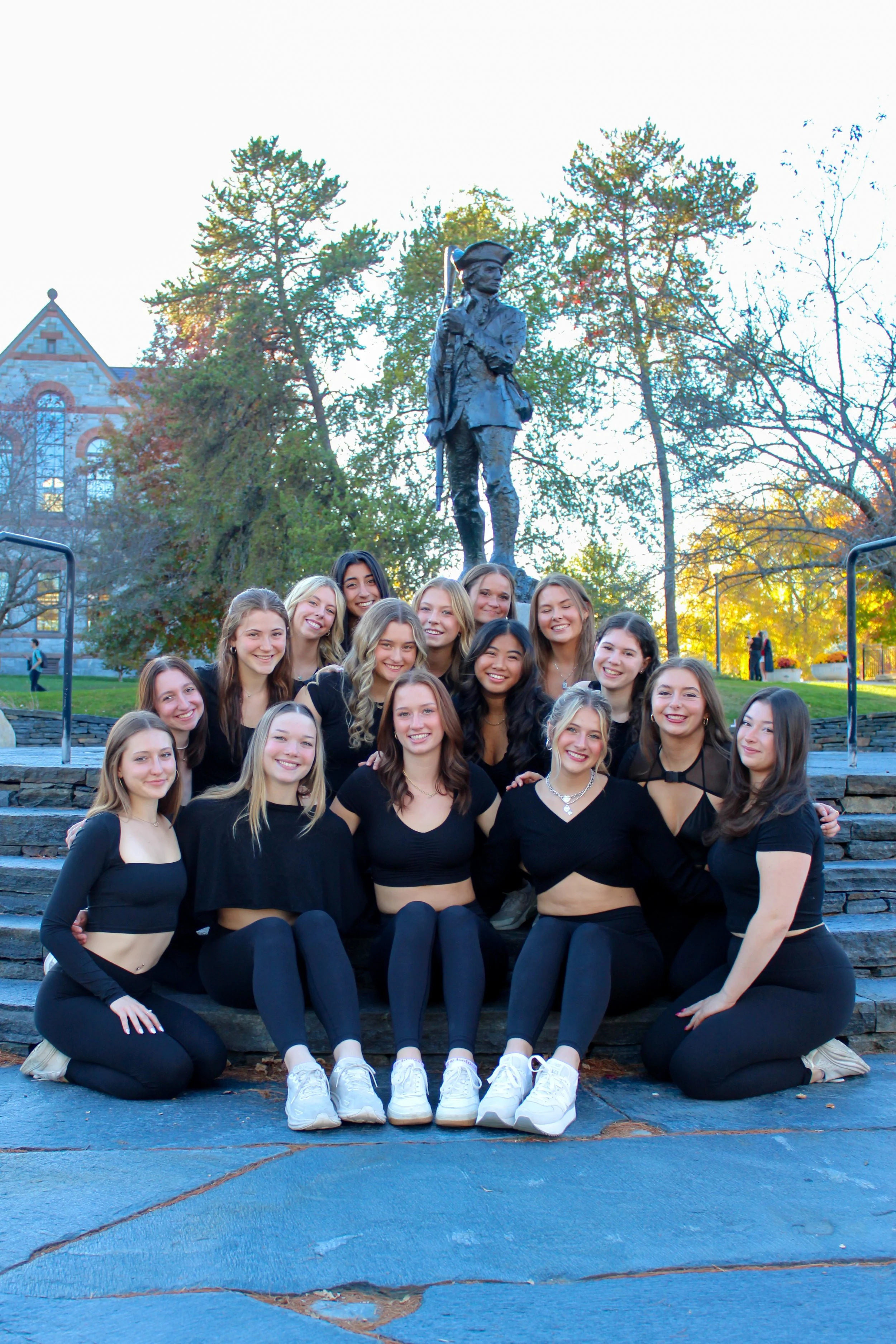 Group of young women wearing black outfits, posing outdoors on steps in front of a statue of a man in historical clothing, trees, and a building in the background, during daytime.