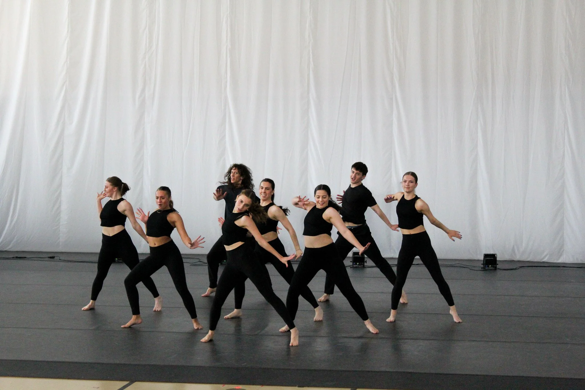 A group of nine dancers in black costumes performing a coordinated dance routine on stage with a plain white backdrop.
