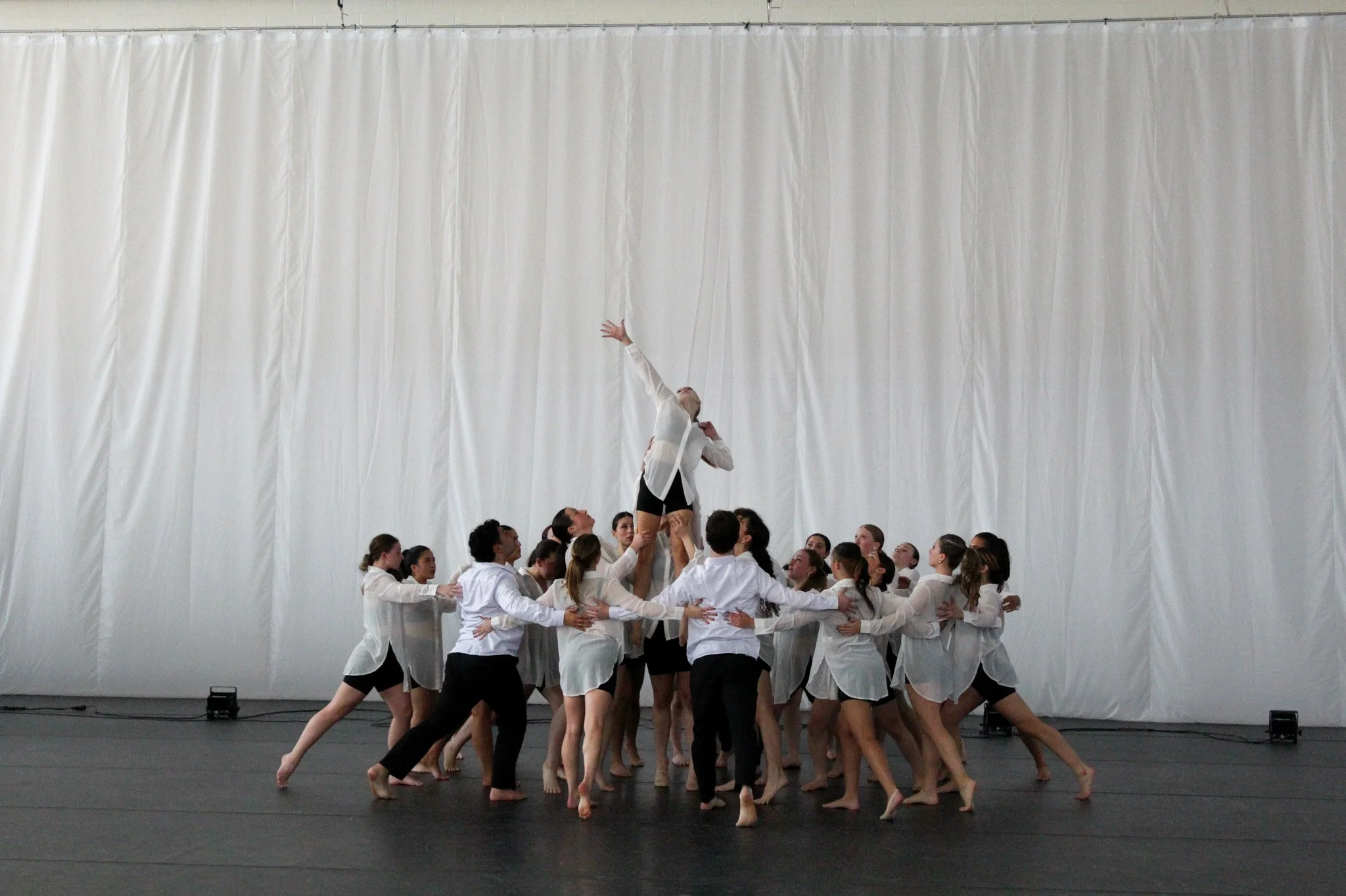 Group of dancers practicing a performance on stage, with one dancer held up by others reaching for the ceiling, against a plain white backdrop.
