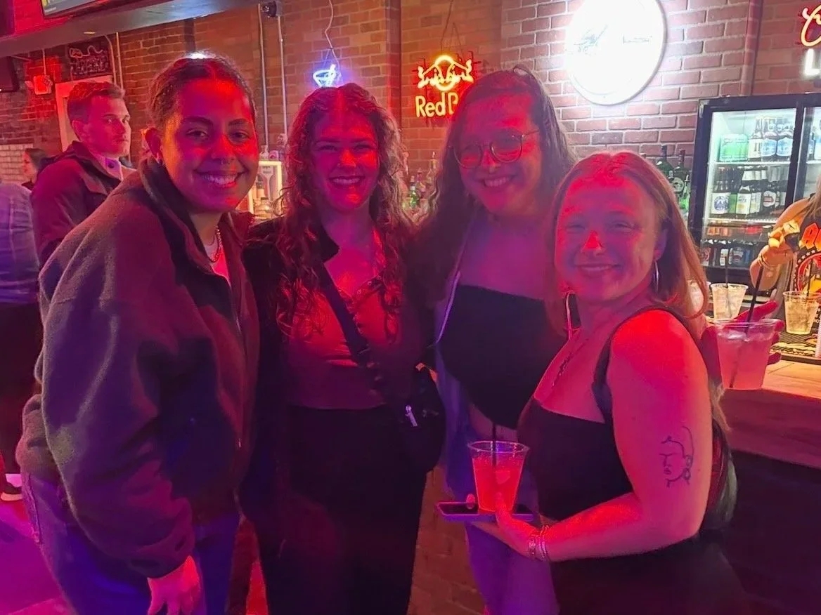 Four women are smiling and posing for a photo in a bar with a brick wall background, neon signs including Red Bull, and drinks on the bar.