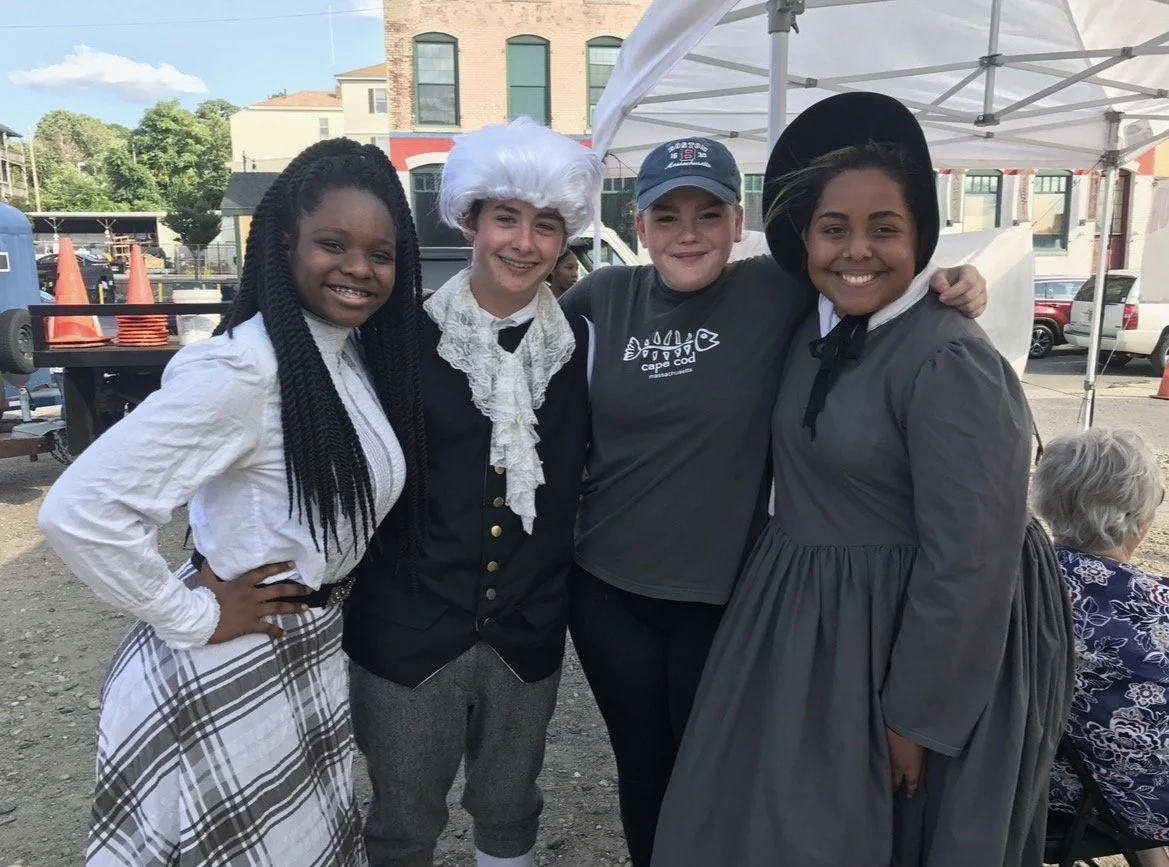 Four women in historical costumes standing outdoors under a white canopy, smiling with their arms around each other.