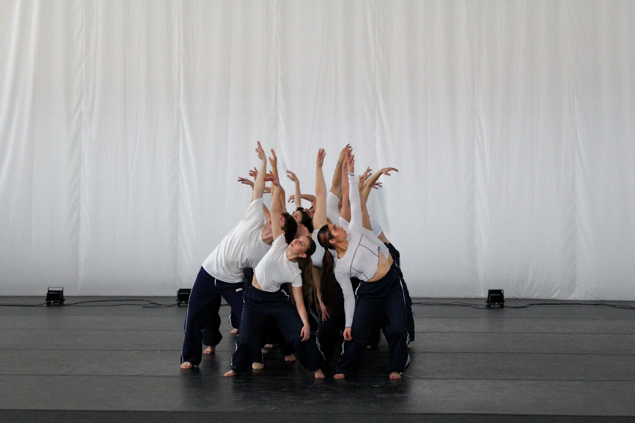A group of dancers is posed in a formation on a stage, with some standing and others kneeling or bending. They are dressed in white shirts and dark pants, with arms extended and heads tilted. The background features a plain white curtain.