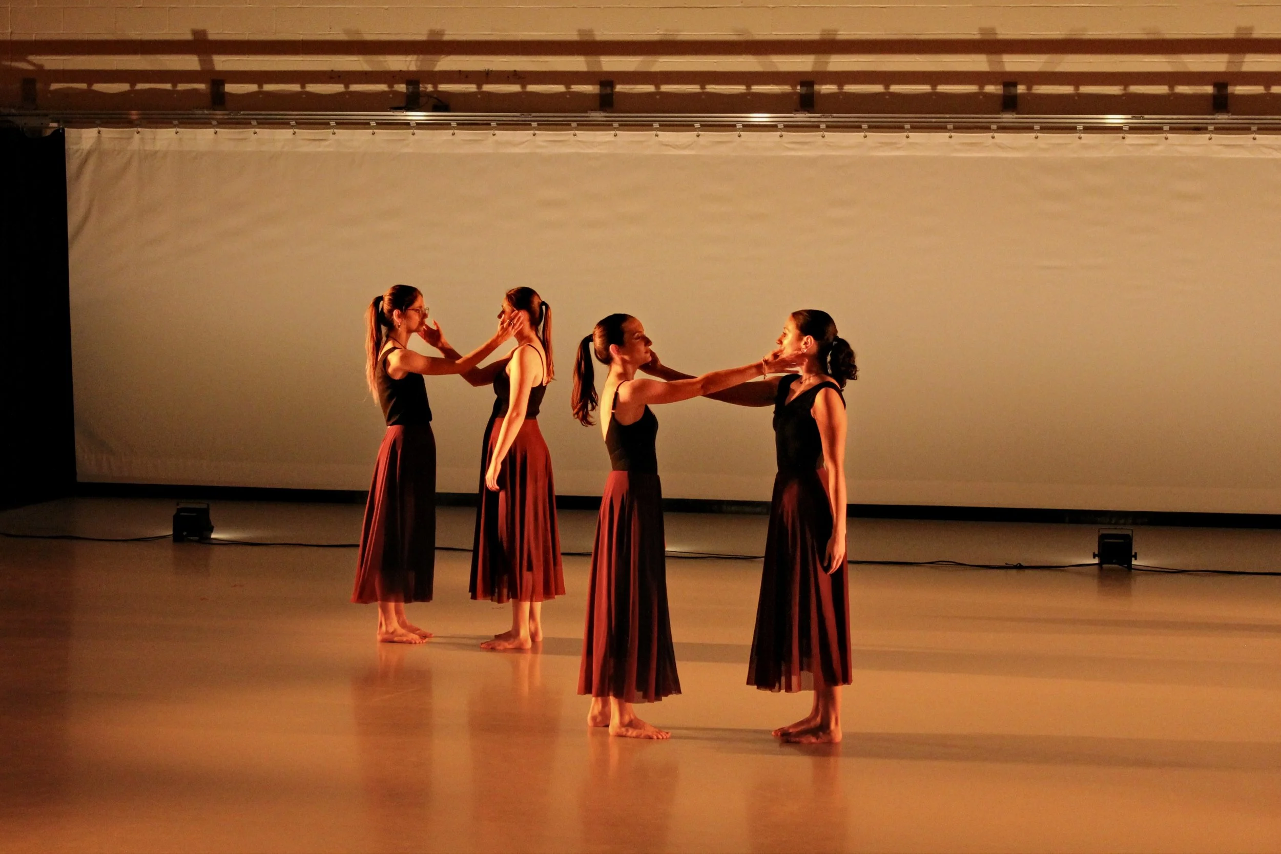 Five women dressed in black tops and long red skirts standing in pairs on a stage, touching each other's faces or necks in a dance performance, with a plain background.