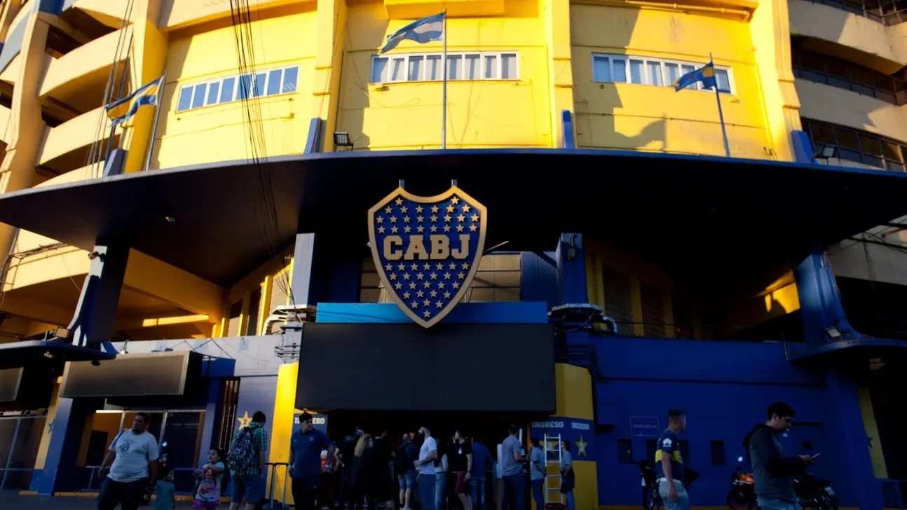 Exterior of a stadium painted in yellow and blue with a shield-shaped sign displaying CABJ and surrounded by small stars. People are entering and exiting the stadium.