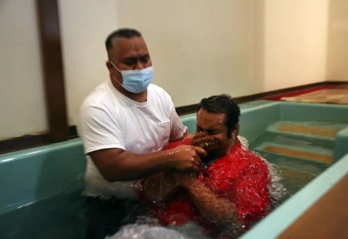 A person in a white shirt and face mask helping another person who is crying and covered in water during a baptism ceremony.