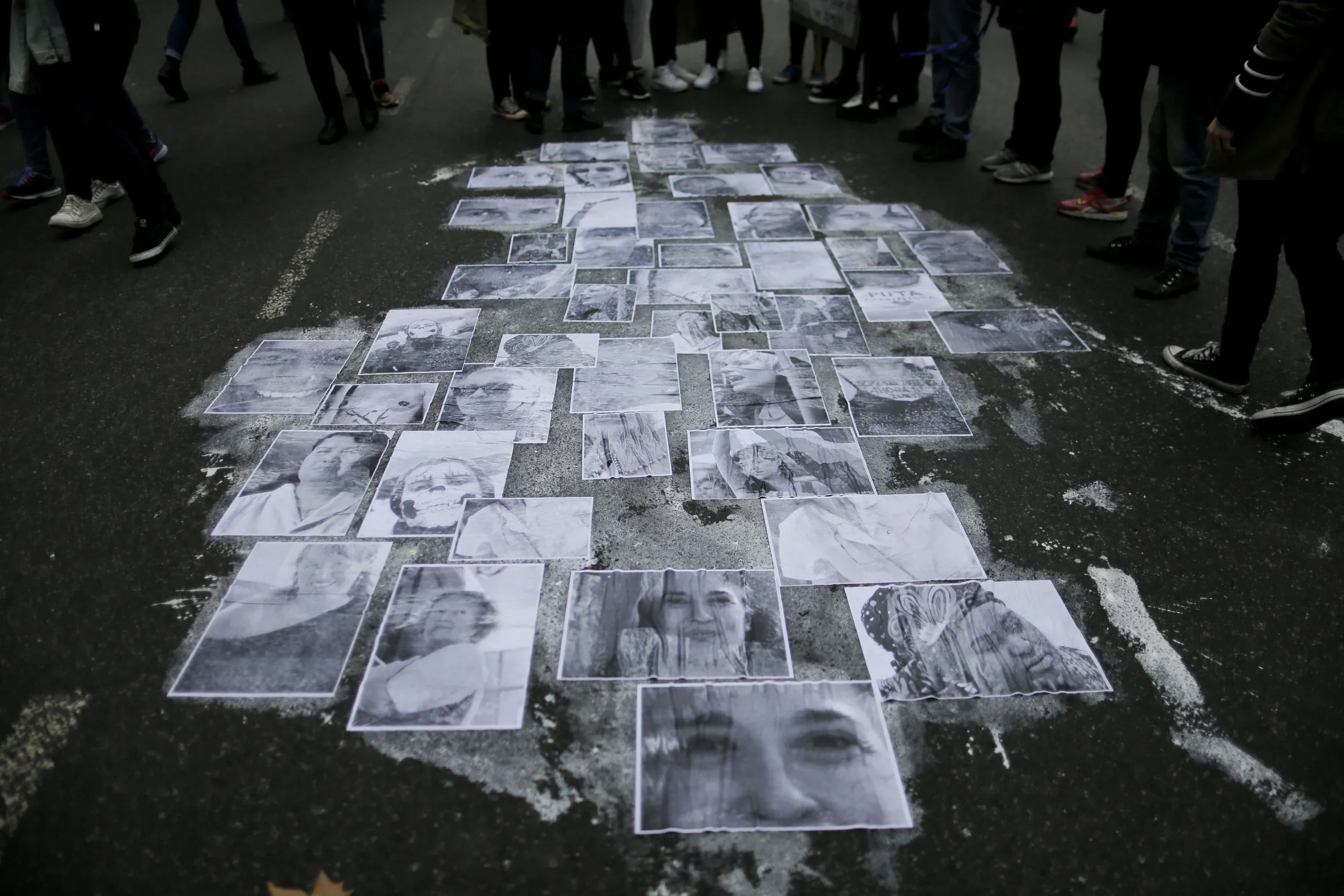 Black and white photographs of people's faces and figures are laid out on a dark street, surrounded by a crowd of people standing around and observing.