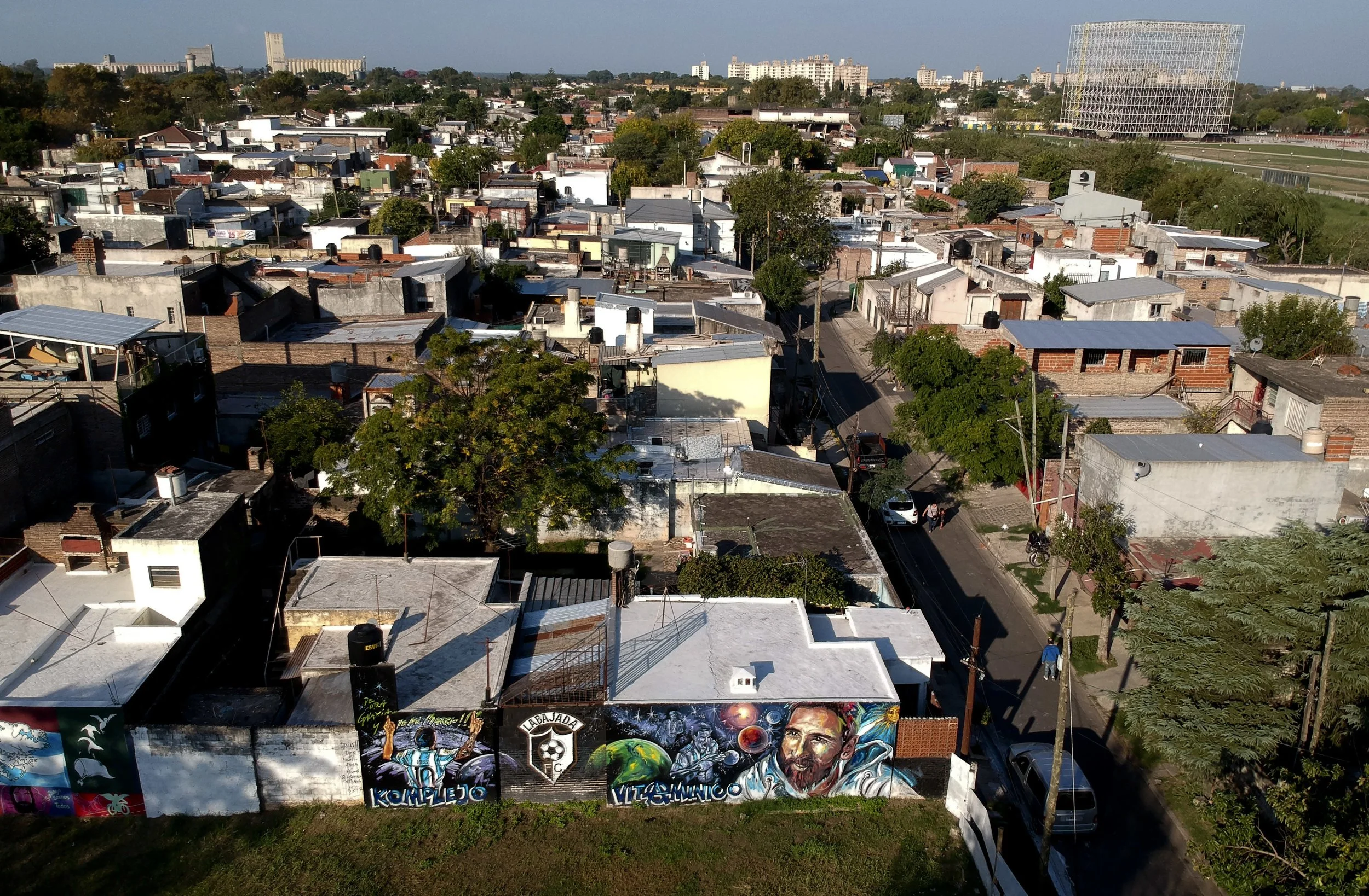 Aerial view of a neighborhood with residential houses, some with metal roofs, and a mural on a wall featuring a soccer player and a smiling man, with trees lining the streets and a large grid-like building in the background.