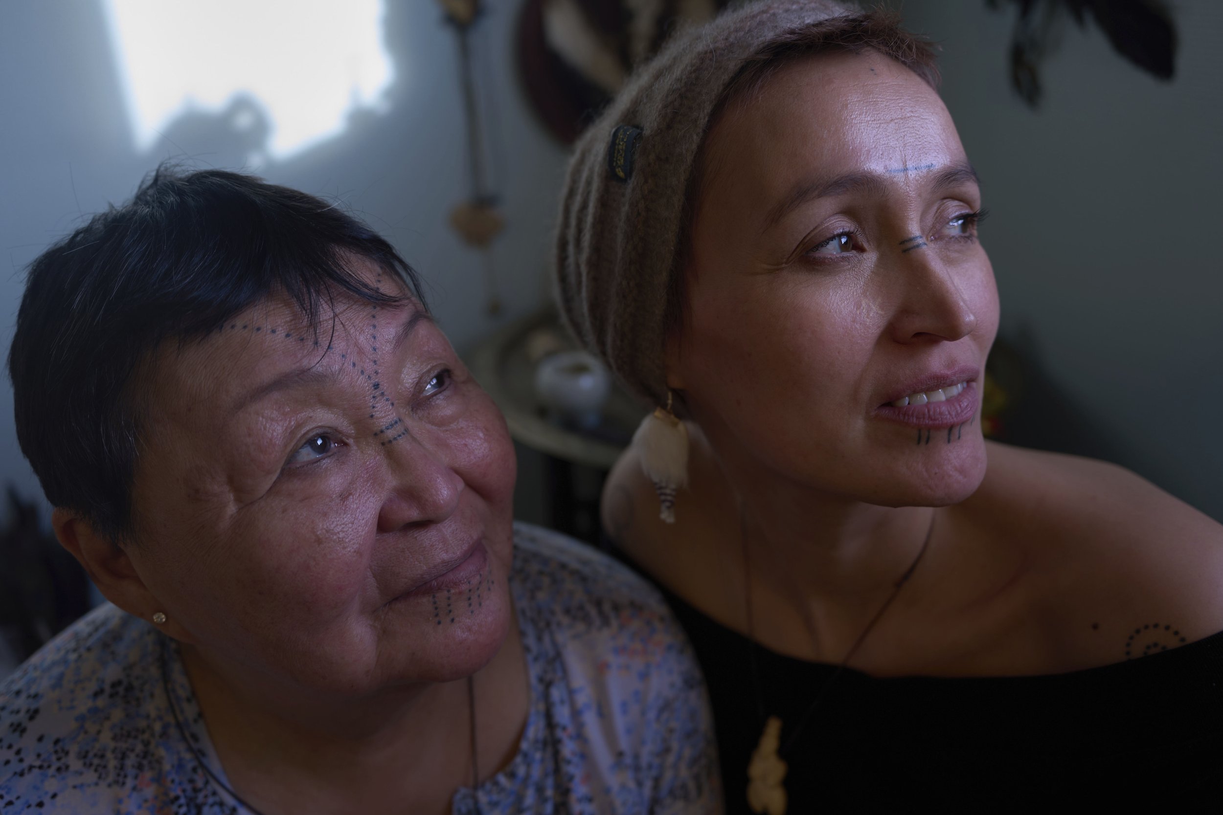 Two women with traditional face paint and jewelry, looking upwards in a close-up shot.