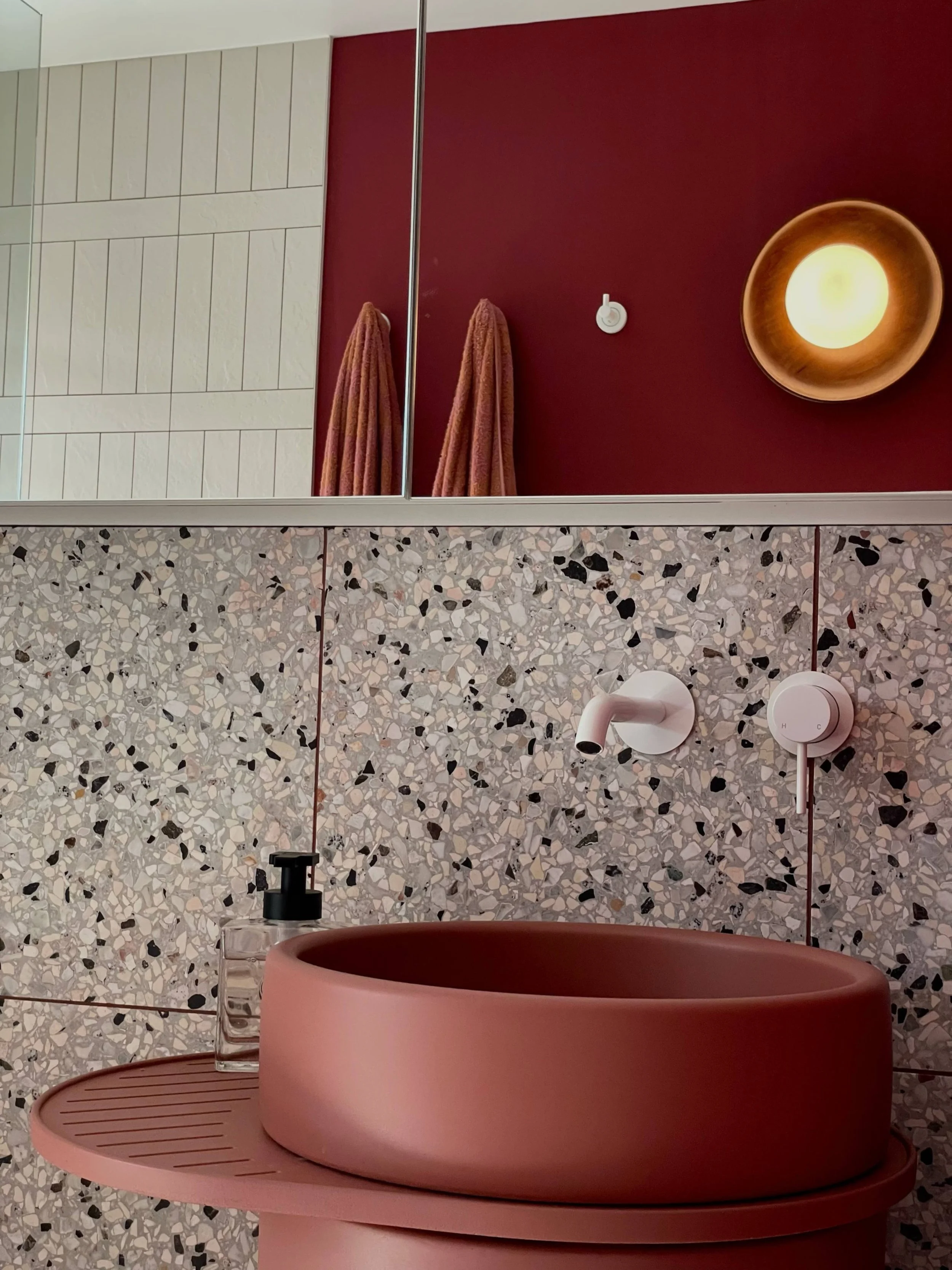 Wall mounted light in a modern bathroom, with burgundy painted walls, terrazzo tile and white vertical tile, burgundy wash basin, white tap and hand soap. Light has been installed by Carfax Connections, a local Sunshine Coast electrician.