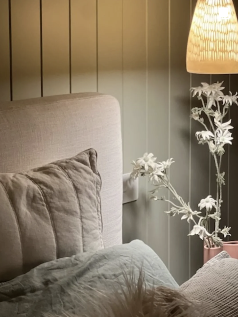 Modern bedroom with green VJ panelling behind neutral headboard, with linen cushions, glowing pendant light and flowers. Light installed by Carfax Connections, Sunshine Coast Electricians