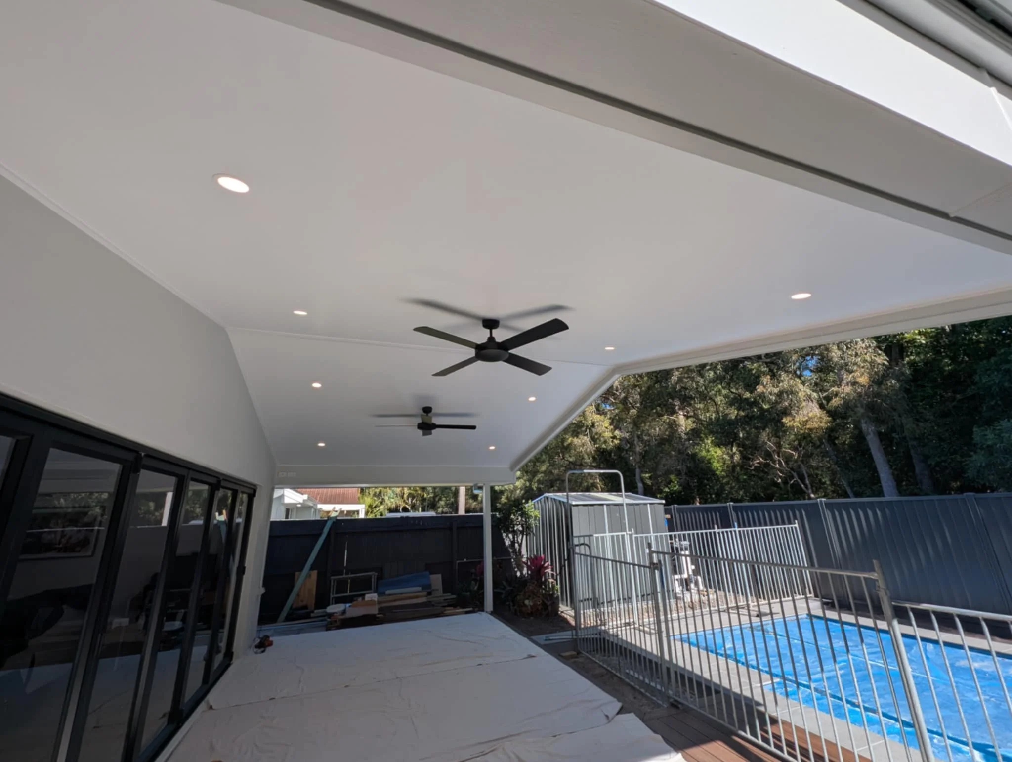 Two black ceiling fans and seven downlight above patio area of modern house, with black framed bifold doors and a swimming pool with a blue cover and white railings. 