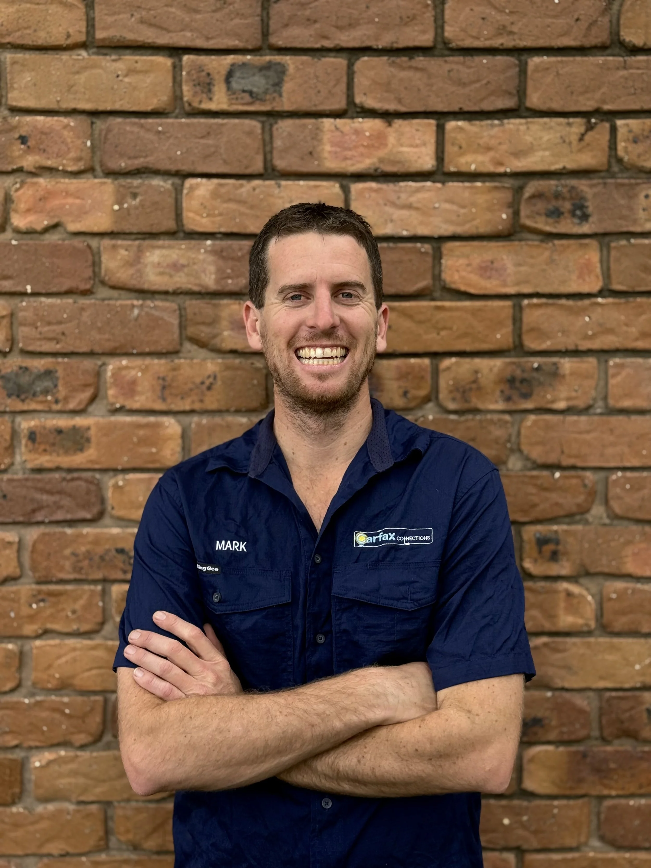 Mark McAleese is the owner of Carfax Connections, a local electricians based on the Sunshine Coast, Australia. Mark is wearing electricians uniform, smiling with arms crossed leaning against a brick wall.