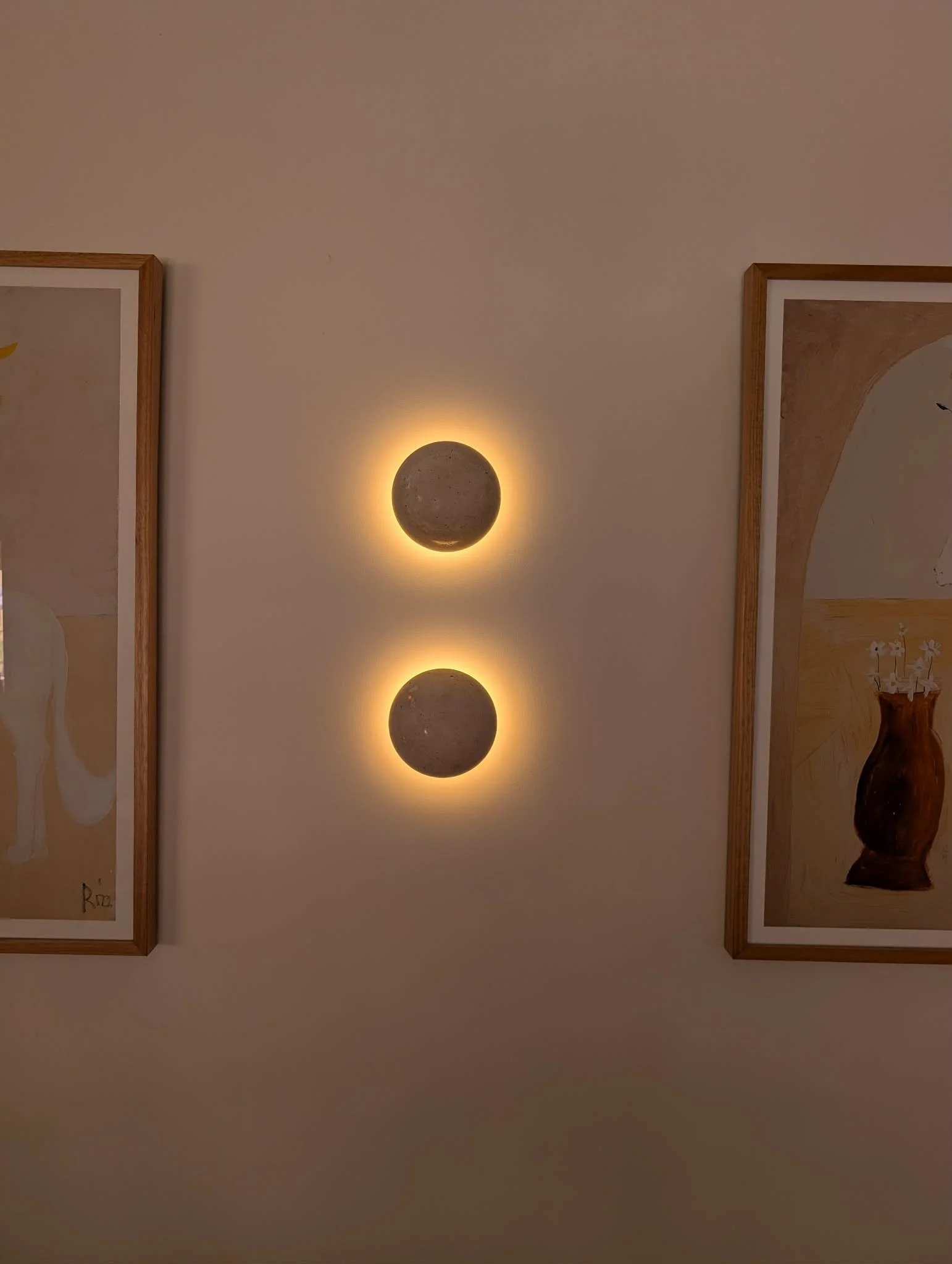 Two round wall mounted lights on a cream wall, between two framed pieces of art in wooden frames