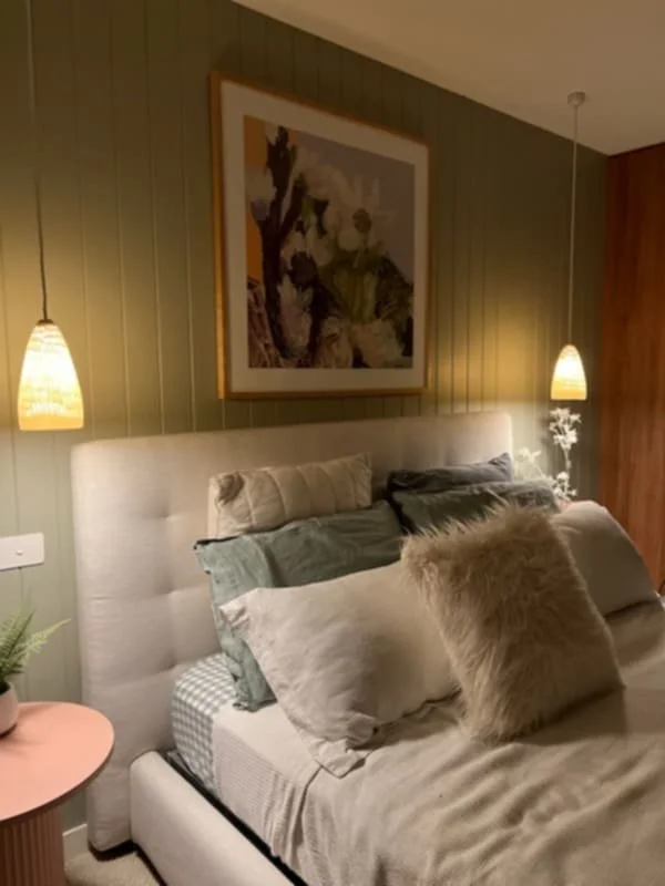 Modern and warm bedroom with neutral decor, green VJ panelling, framed artwork of flowers, red bedside tables, scatter cushions, carved pendant lighting