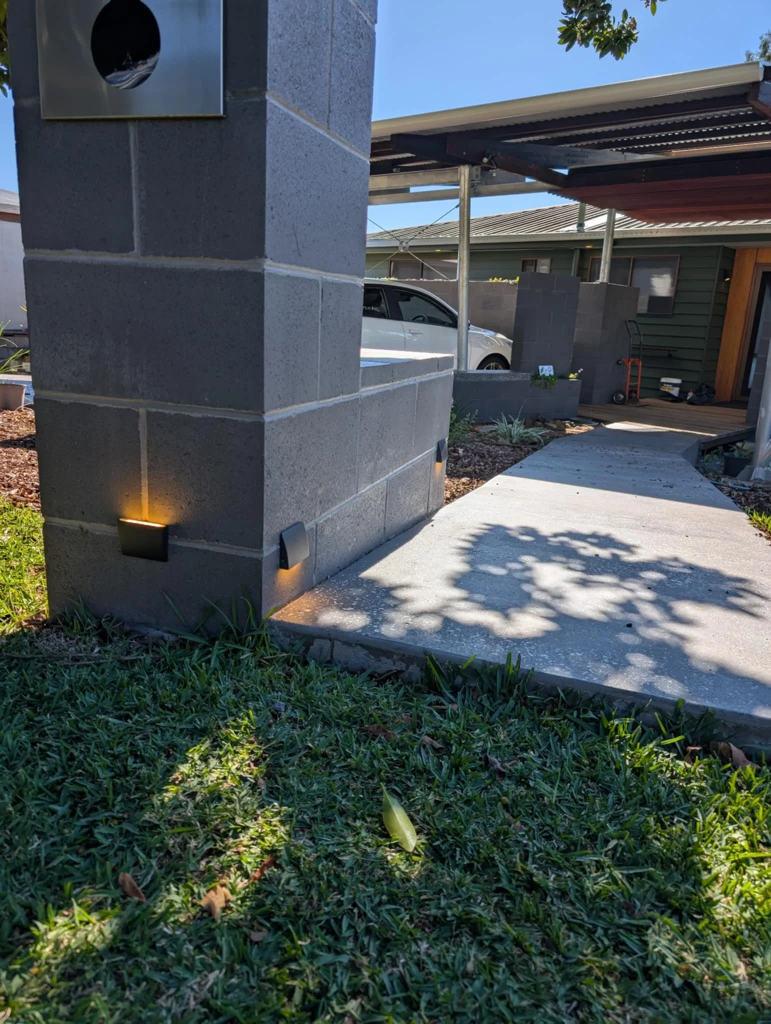 Garden wall mounted lighting on grey brick wall next to concrete driveway. Green grass, front yard leading to house.