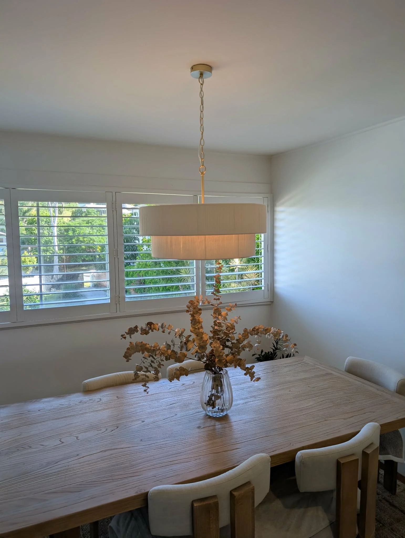 Drop pendant light with natural structured lampshade displaying a glowing light over a wooden dining tablee, wood and fabric dining chairs, white walls, blinds