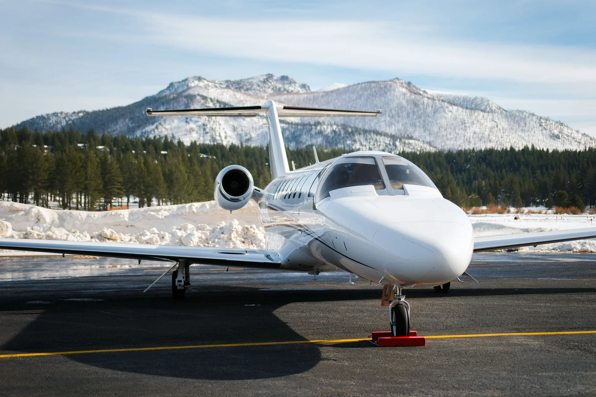 A white private jet parked on a runway with snow-covered mountains and a forest of pine trees in the background.
