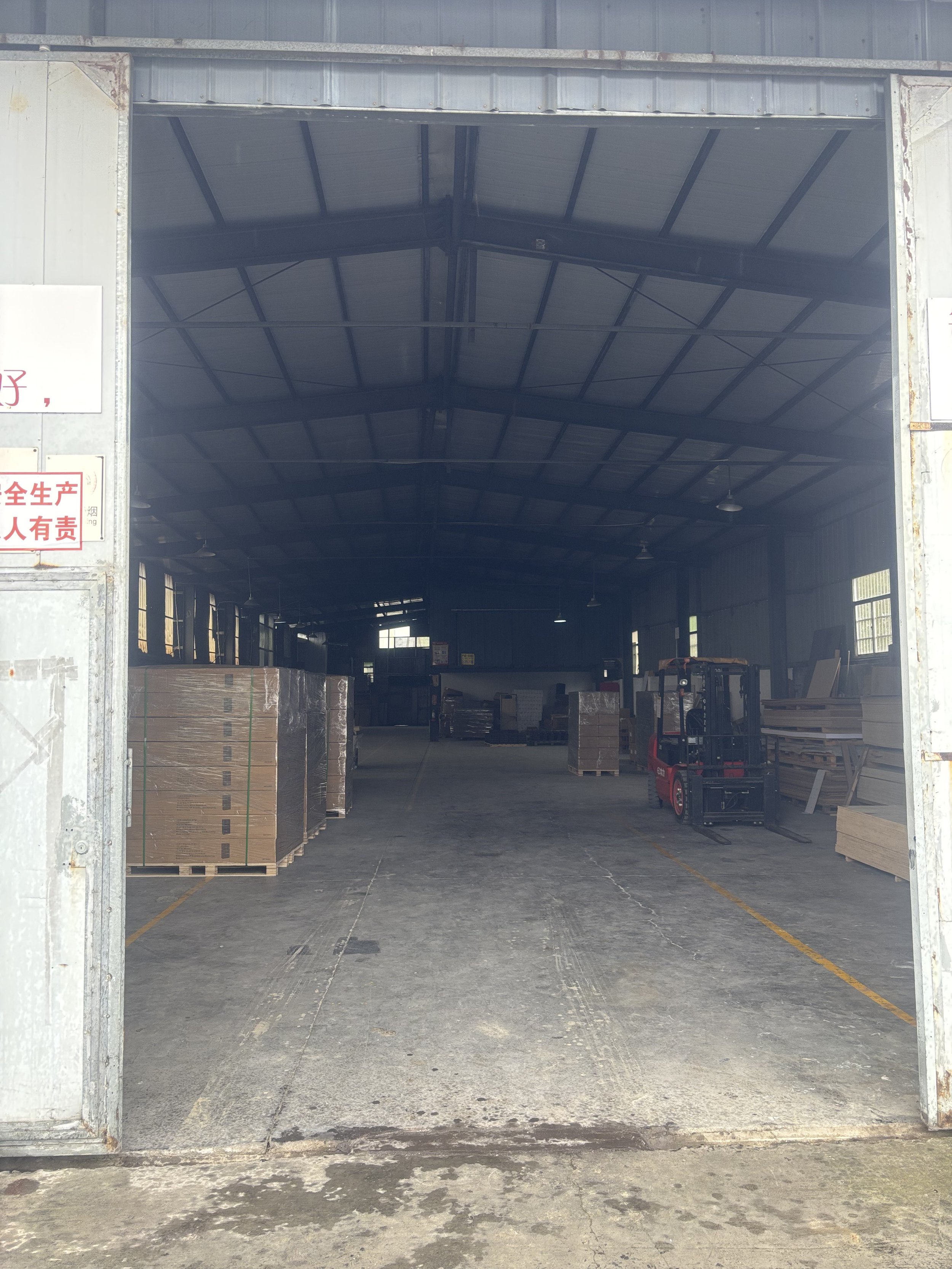 Interior of a warehouse with pallets of materials on the left, a forklift on the right, and stacks of wood on the far right, under a high metal ceiling.