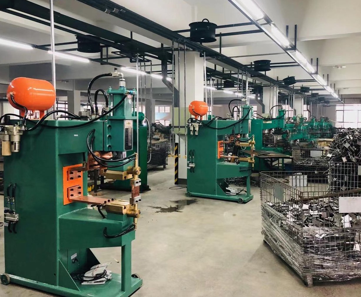 Industrial factory with green machines, orange tanks, and metal scrap baskets in a well-lit warehouse. A group of welding machines for metal products