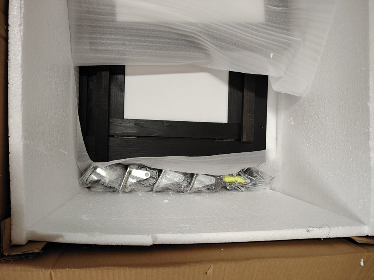 Small black wooden picture frame, wrapped in clear plastic, inside white foam packaging with some yellow and silver hardware tools or parts, on a brown cardboard surface.