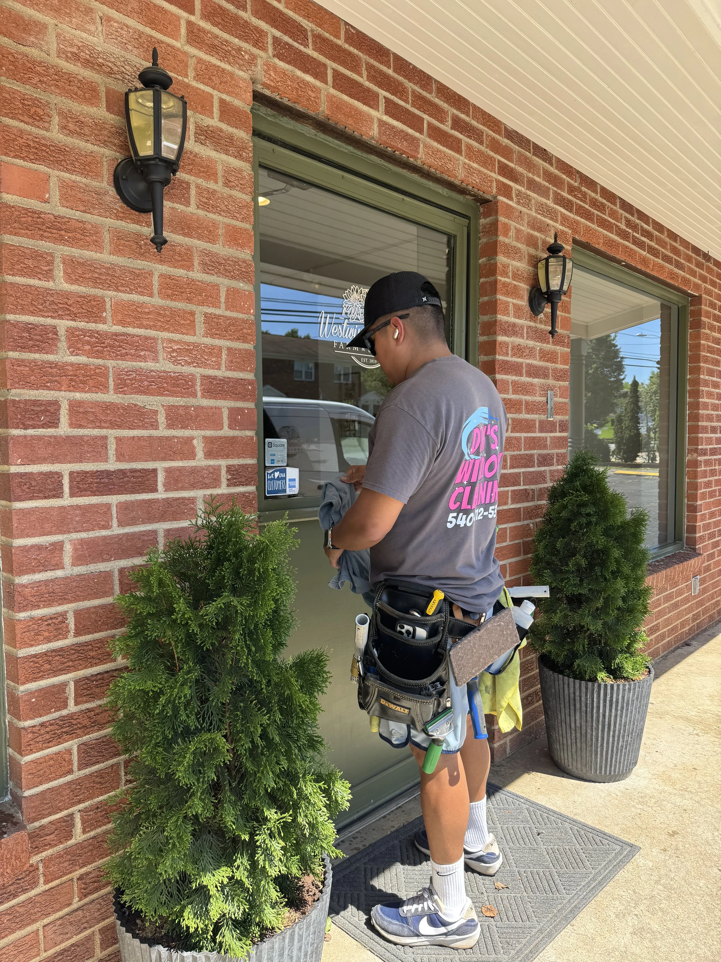 A young man with a tool belt and professional window cleaning tools cleans the glass on the front door of a commercial brick buiding.