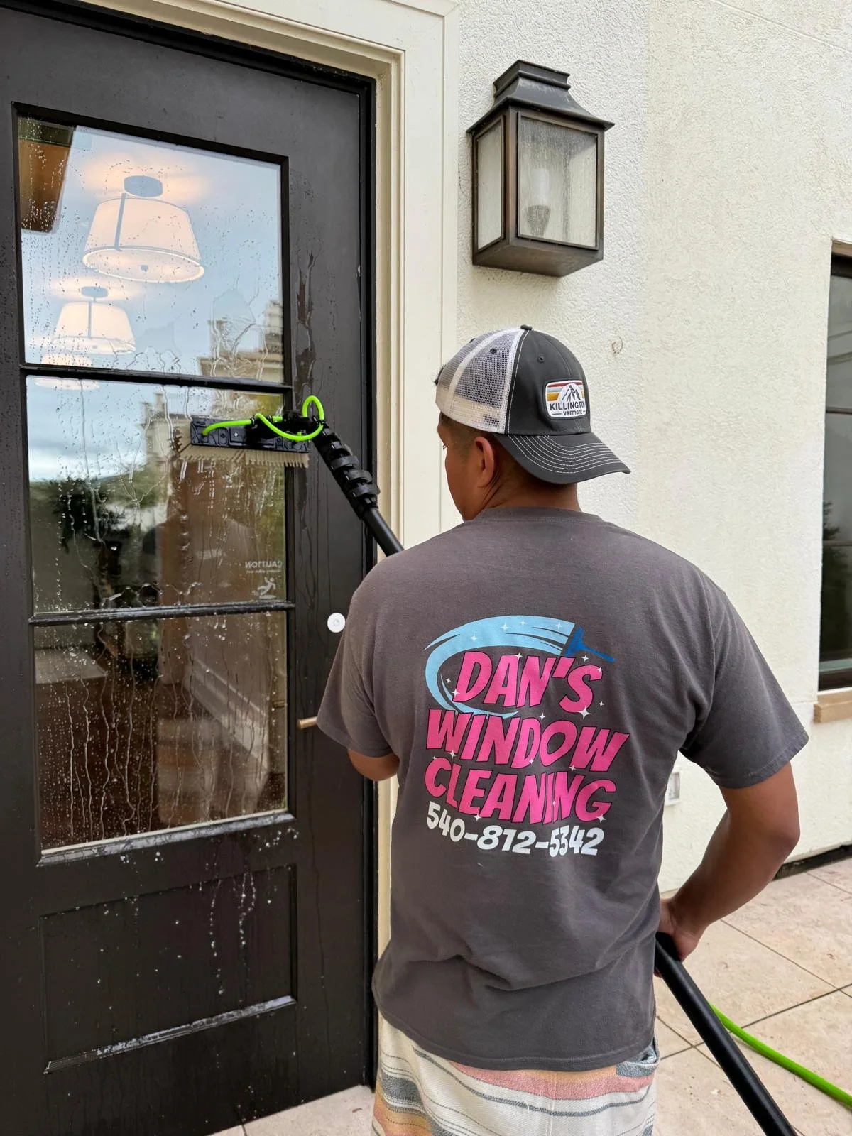 A man wearing a gray t-shirt with the text 'Dan's Window Cleaning' and a phone number, cleaning a glass door with a pressure washer, water splashing on the door, and reflections visible in the wet glass.