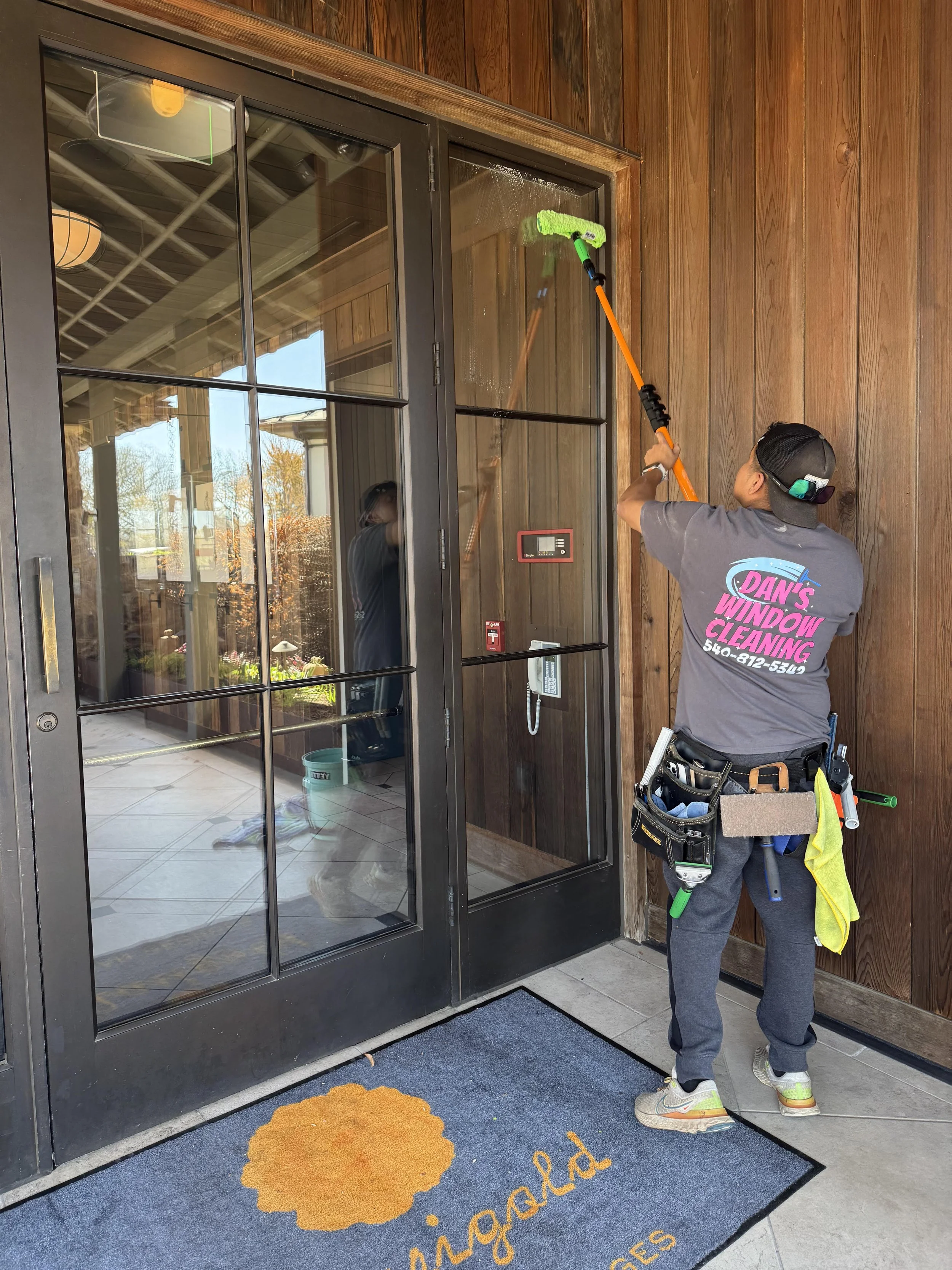 A worker cleaning a glass door with a window squeegee. The worker is wearing a gray shirt with pink and white text that reads 'Dan's Window Cleaning' and a phone number. The worker has a tool belt and is wearing a black cap and sunglasses. There is a