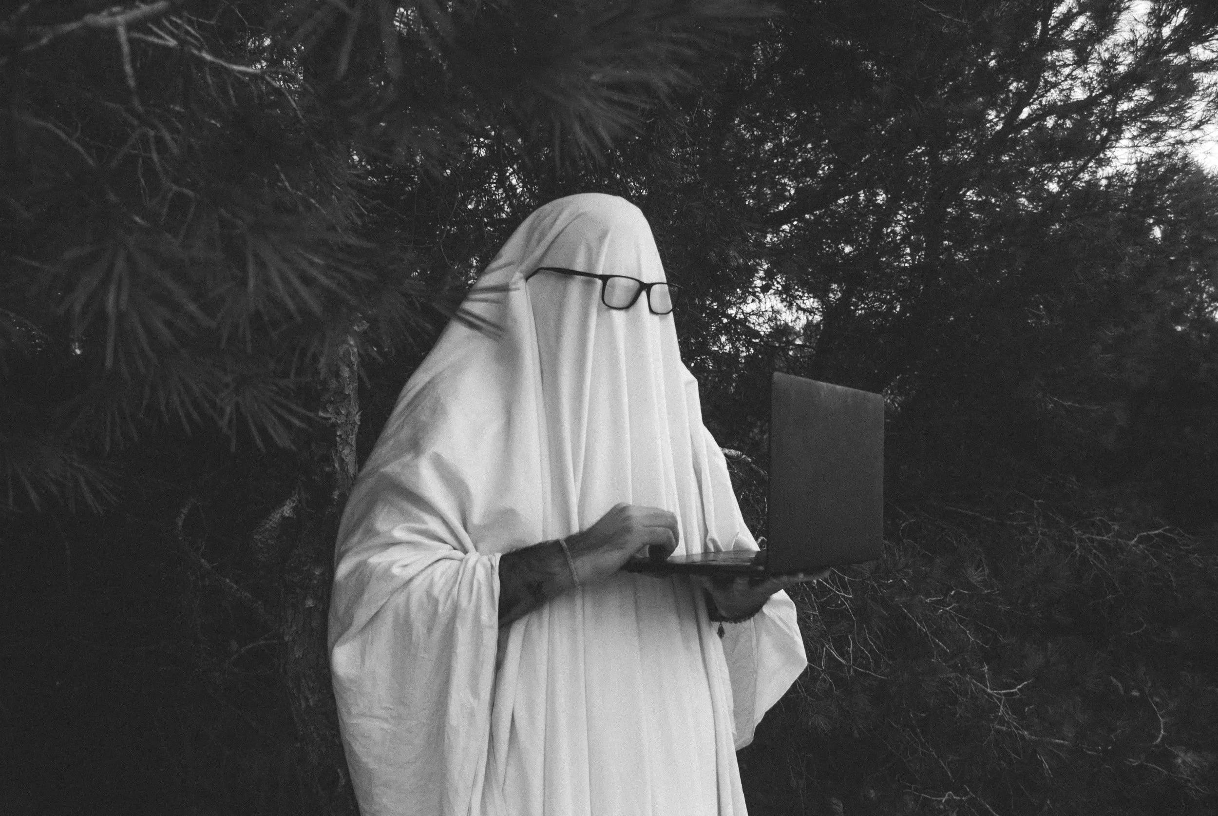 A person dressed as a ghost, wearing glasses, holding a laptop in an outdoor wooded area.