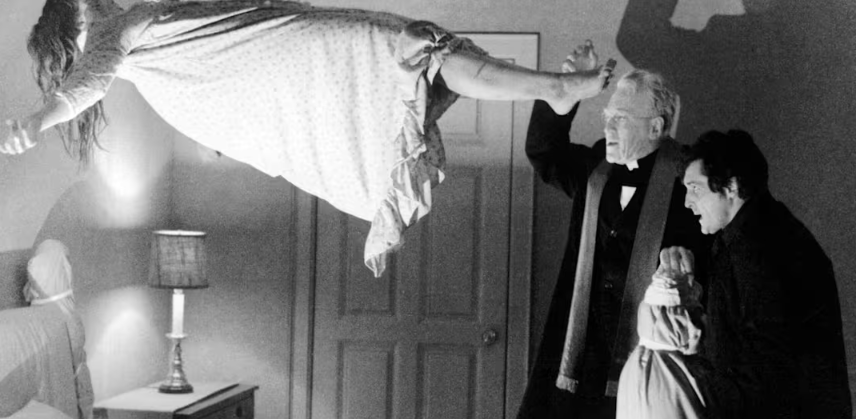 A man with white hair and a woman with dark hair are standing in a bedroom, holding the woman's arm and raising her hand. A girl in a dress is floating horizontally above them, with her arms extended.