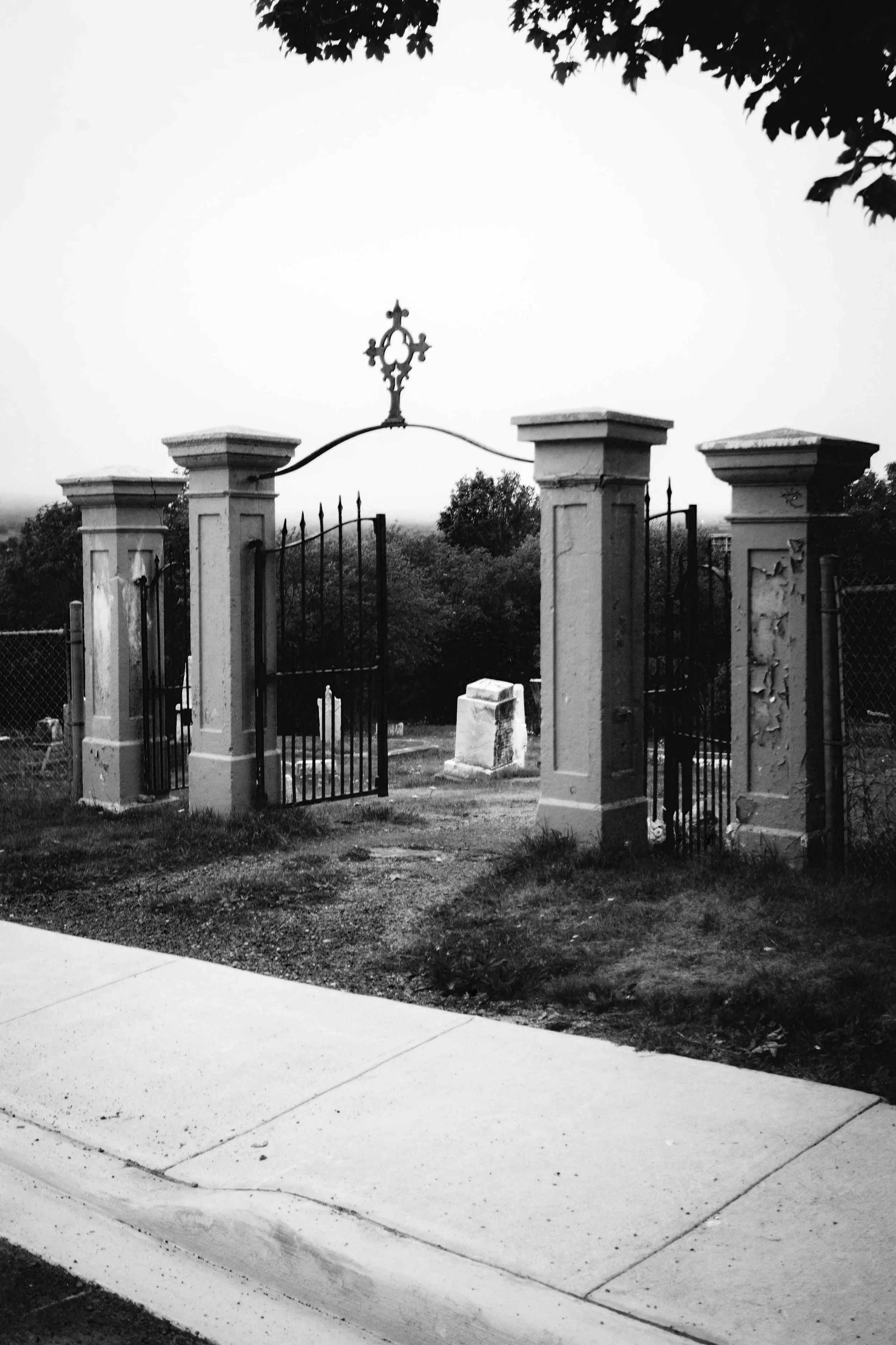 Black and white photo of an old, wrought iron gate with four stone pillars, leading to a cemetery or garden, with trees and a distant headstone visible behind the gate.