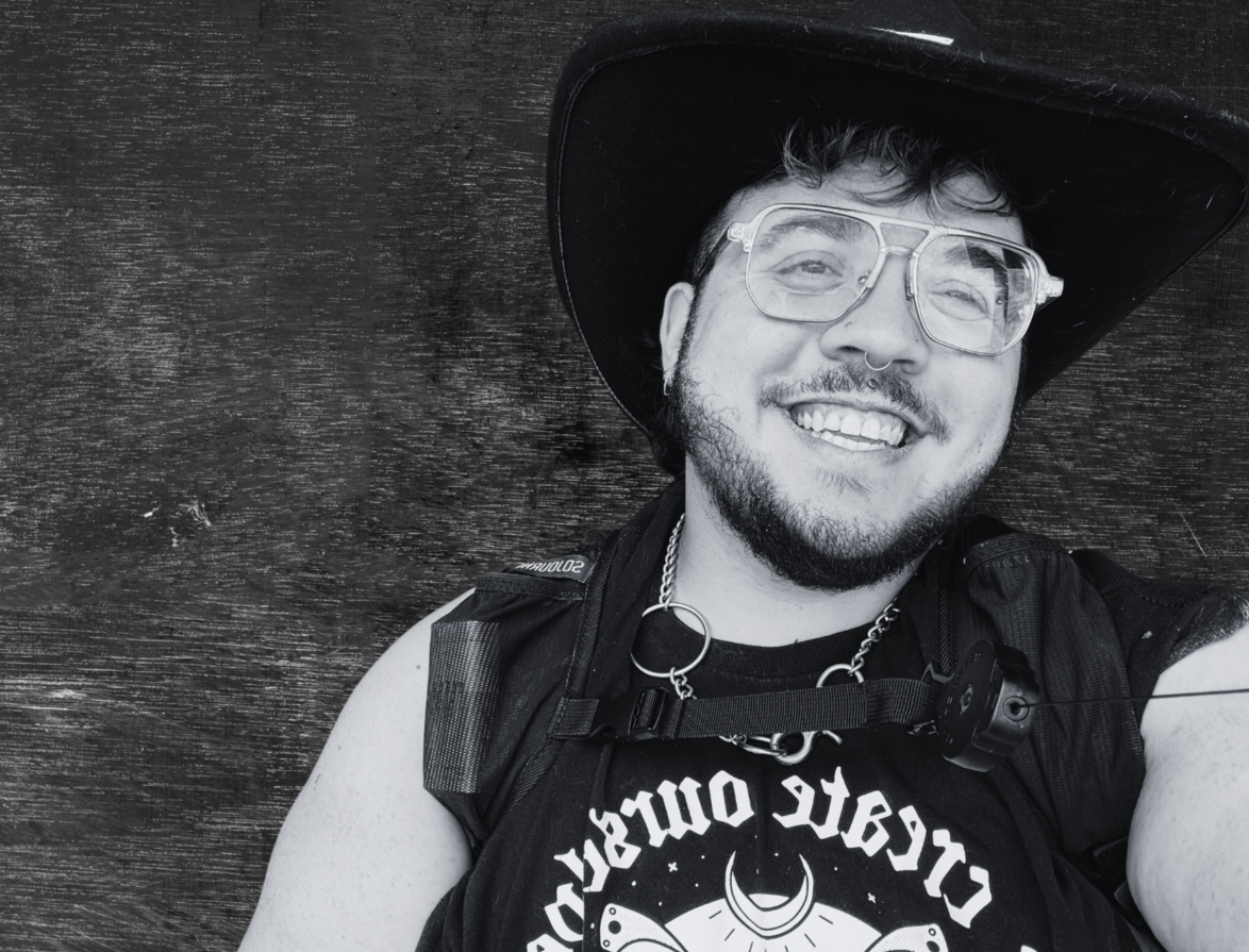 A smiling person with a beard, wearing glasses, a wide-brimmed hat, a black shirt with white text, a backpack, and accessories, lying on a wooden surface.