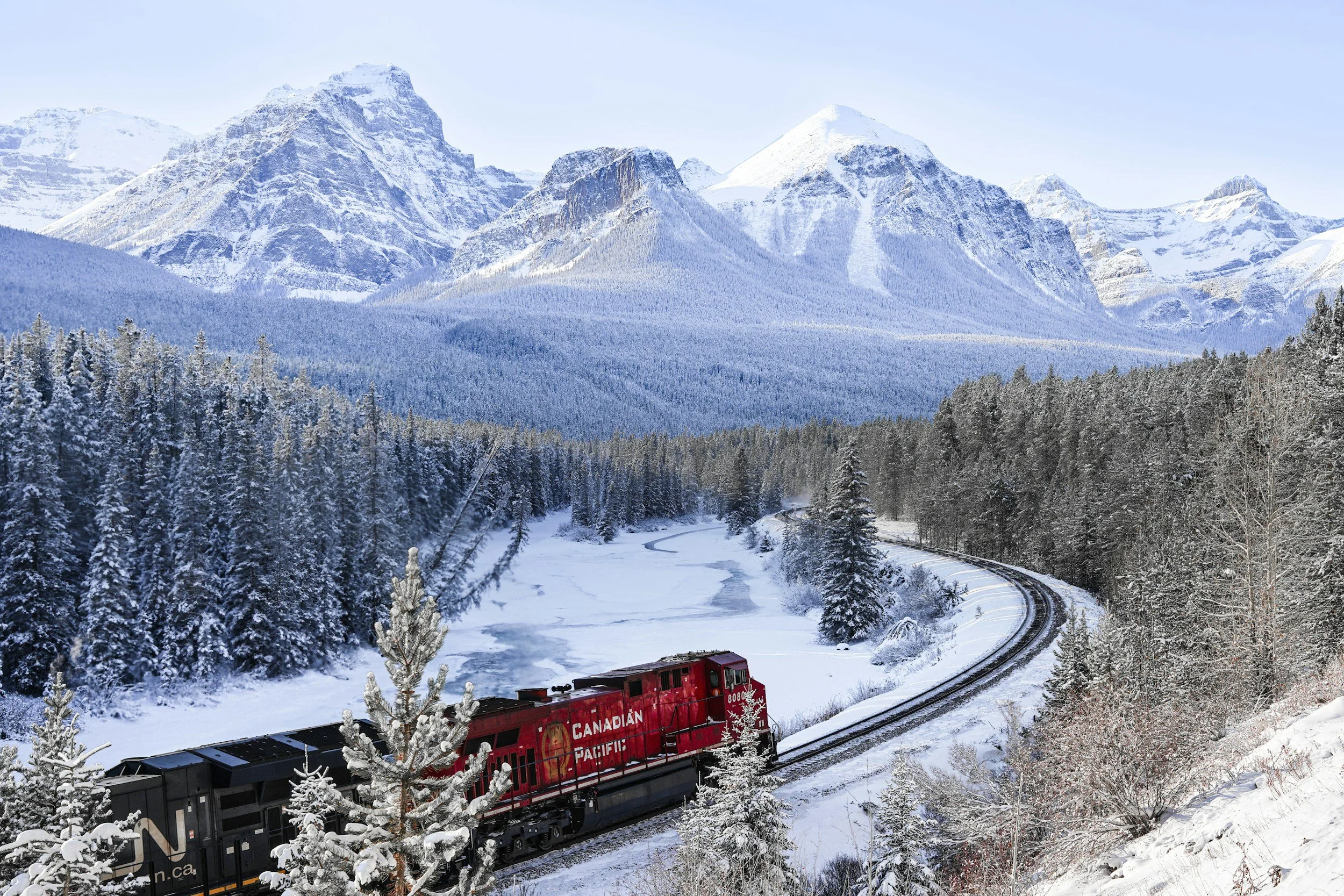 Snow-covered mountains with a forest at their base, a snow-covered traintracked landscape, and a red Canadian Pacific train moving through the snowy terrain.