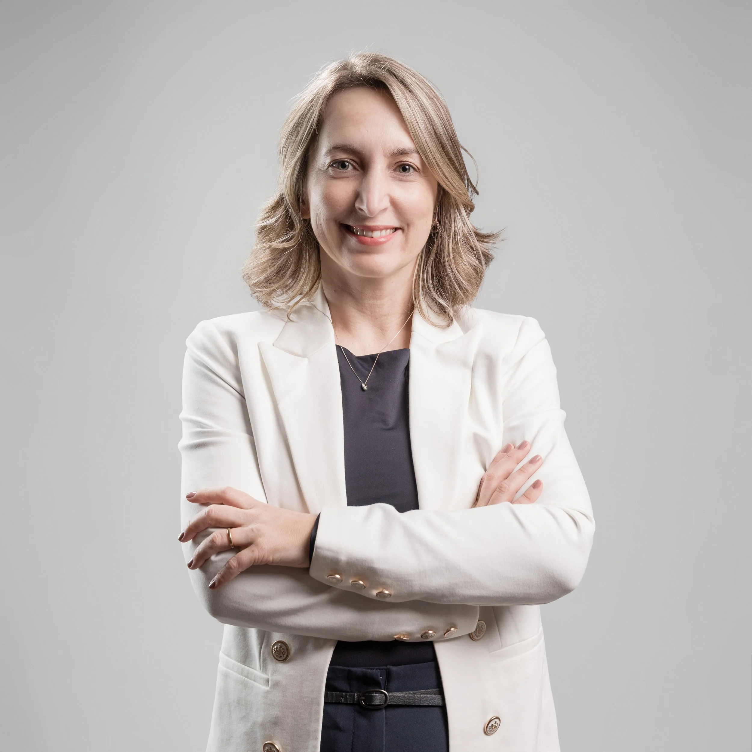 Uma mulher vestindo um blazer branco sobre uma blusa preta, em pé com os braços cruzados, contra um fundo liso cinza claro.