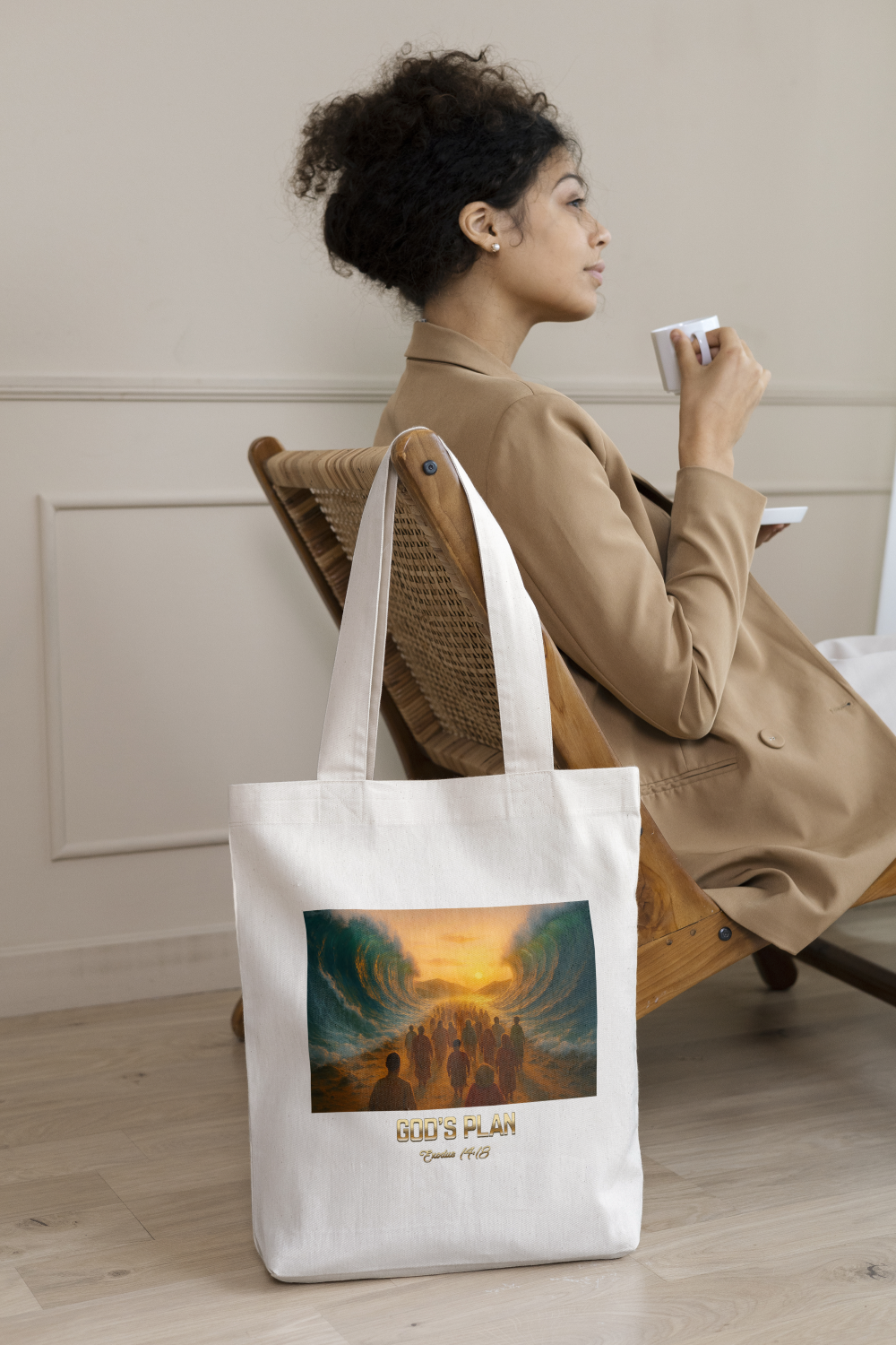 Woman sitting in a wooden chair, drinking from a white cup, with a white tote bag featuring an artistic image and the words "GOD'S PLAN" and "Ephesians 1:16" in front of her.