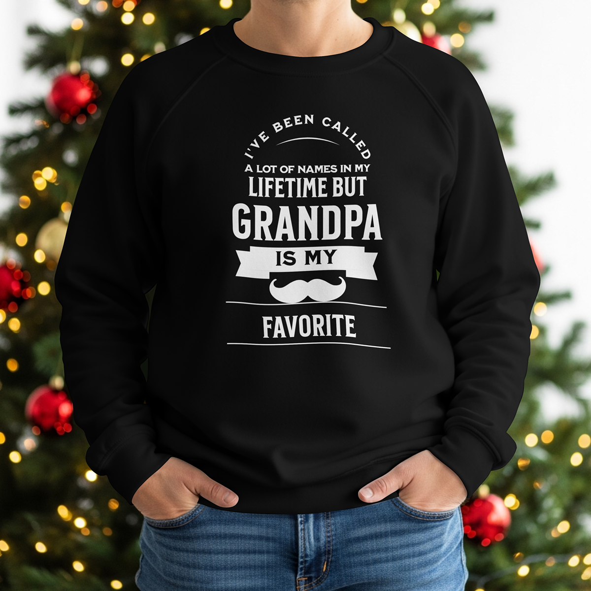 A person wearing a black sweatshirt with bold white lettering and a mustache graphic sta in front of a warmly lit Christmas tree. The sweatshirt reads, “I’ve been called a lot of names in my lifetime, but Grandpa is my favorite