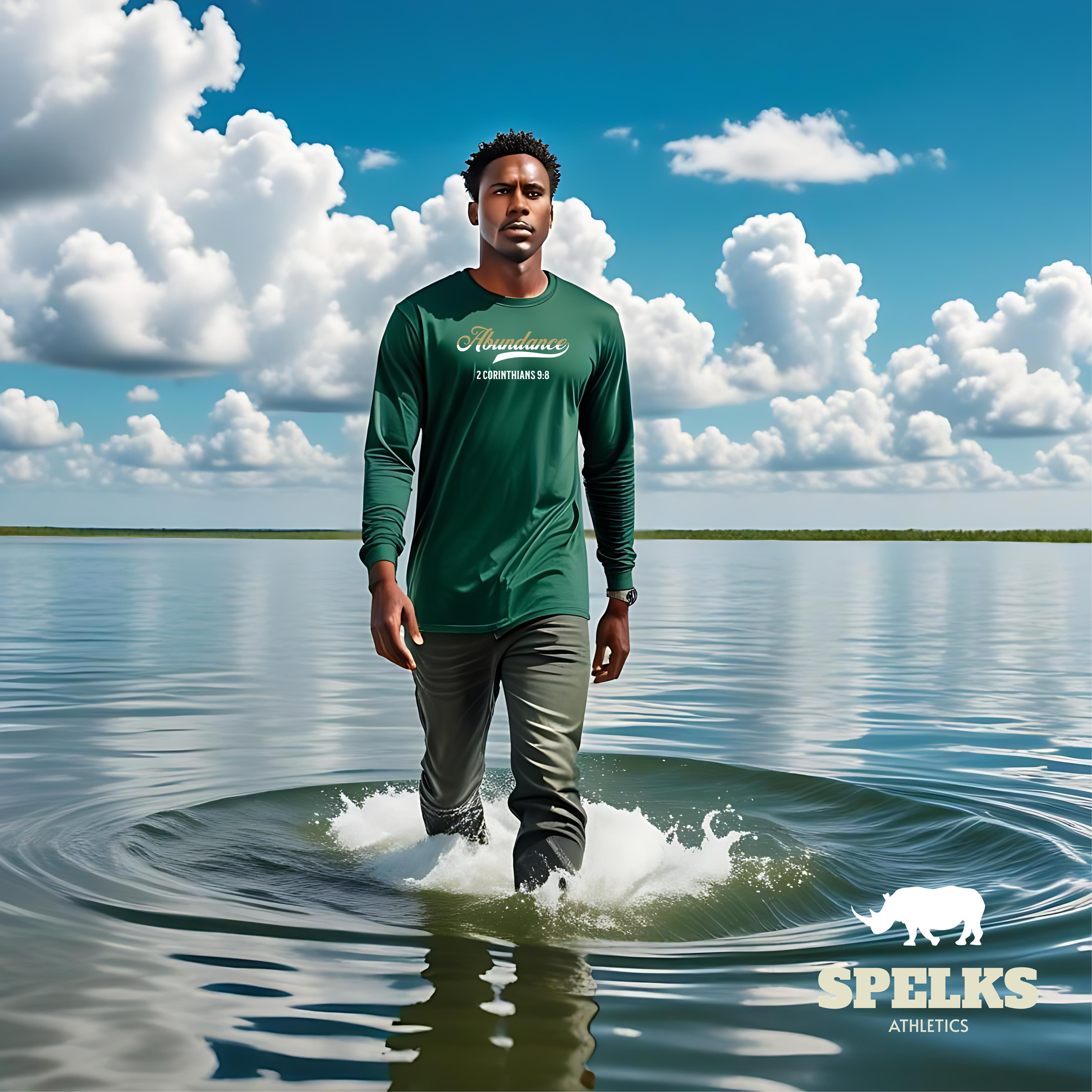 A young man walking in water with a cloud-filled sky in the background. He is wearing a green shirt with the words "Abundance" and a Bible verse reference, and gray pants.