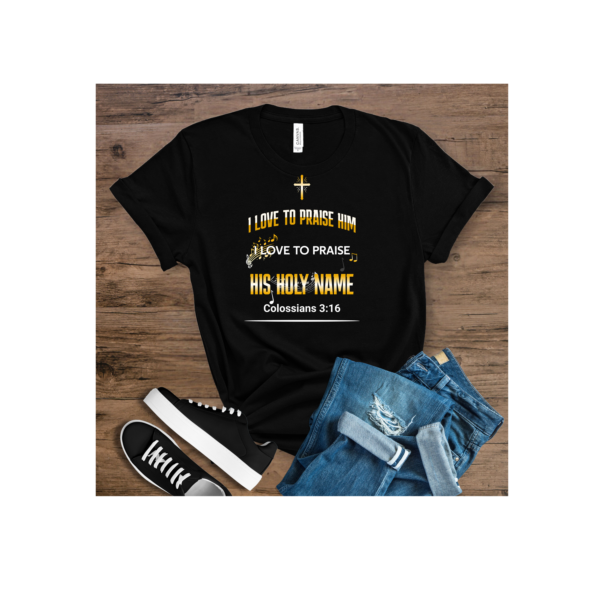 Black T-shirt with religious text and symbols, including a cross and musical notes, laying on a wooden surface with distressed jeans and black sneakers beside it.