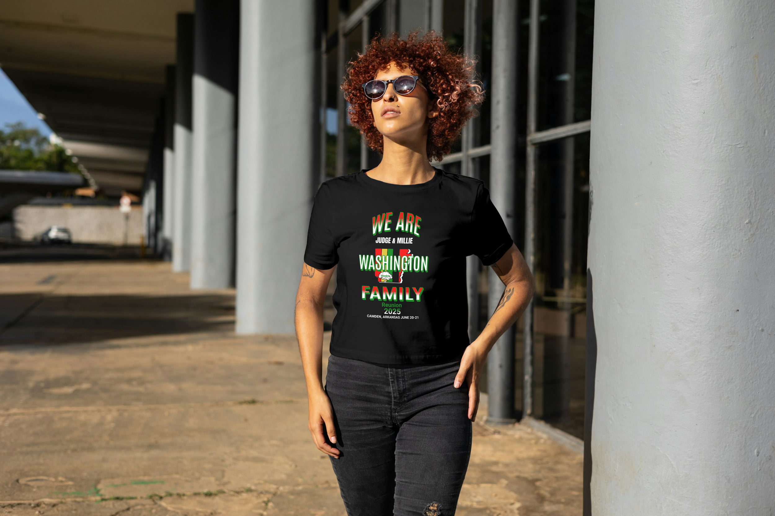 A woman with curly red hair, wearing sunglasses, a black T-shirt with colorful text and graphics, and black jeans, standing outdoors near a modern building with large windows and white columns.