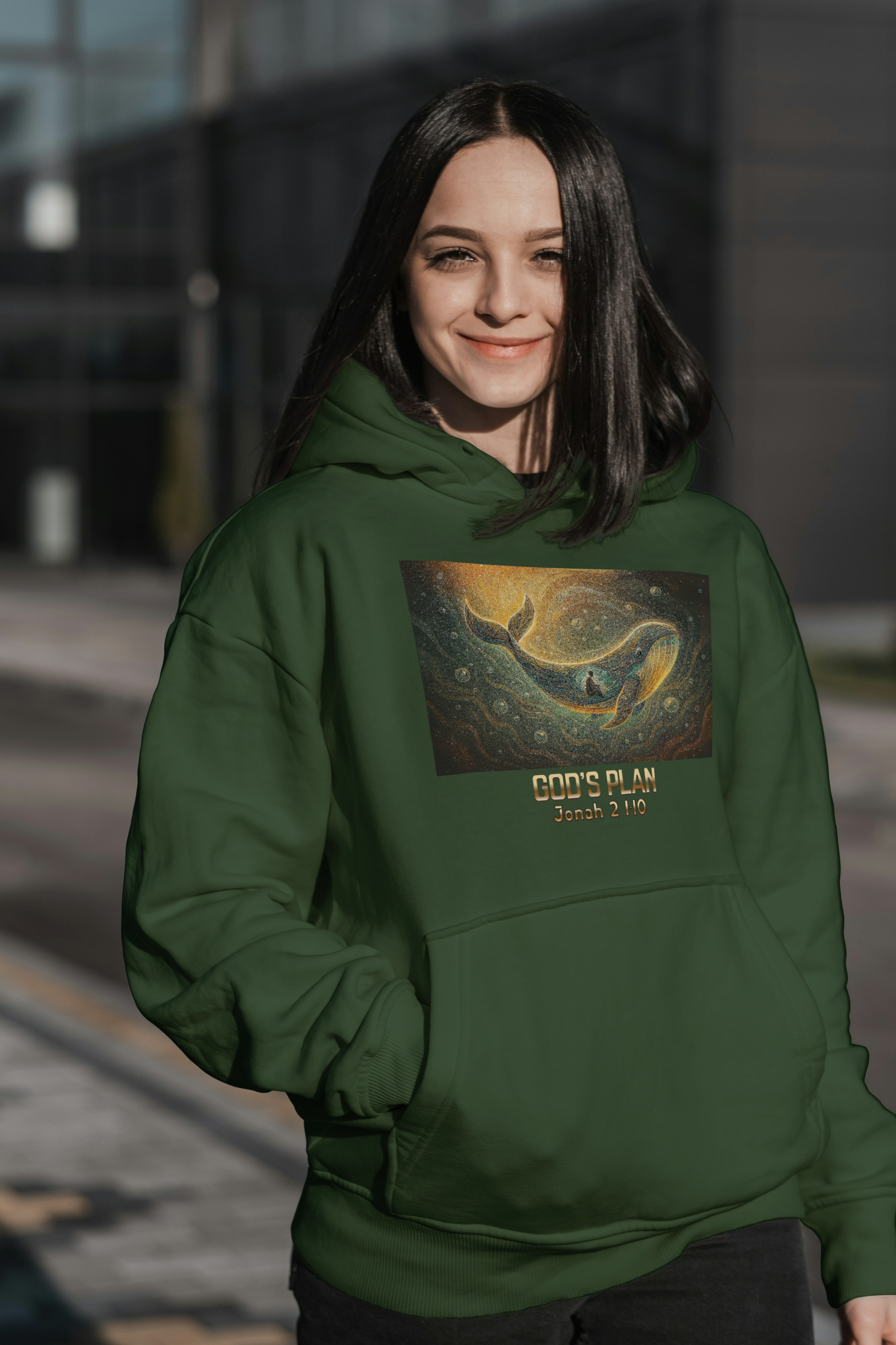 A young woman with black hair wearing a green hoodie that features an artistic depiction of a whale in a galaxy with the text 'GOD'S PLAN Jonah 2:10' on it.