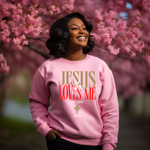 A smiling woman wearing a pink sweatshirt with the text 'Jesus Loves Me' standing outdoors among cherry blossom trees.