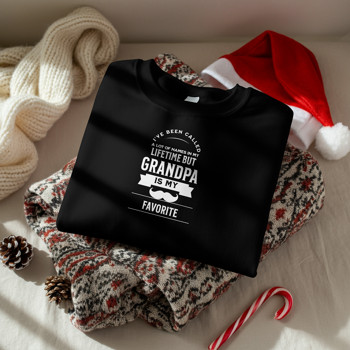 A black T-shirt with a festive Christmas message resting on a patterned holiday blanket, surrounded by a red and white Santa hat, a cream-colored knitted scarf, pine cones, and a candy cane.