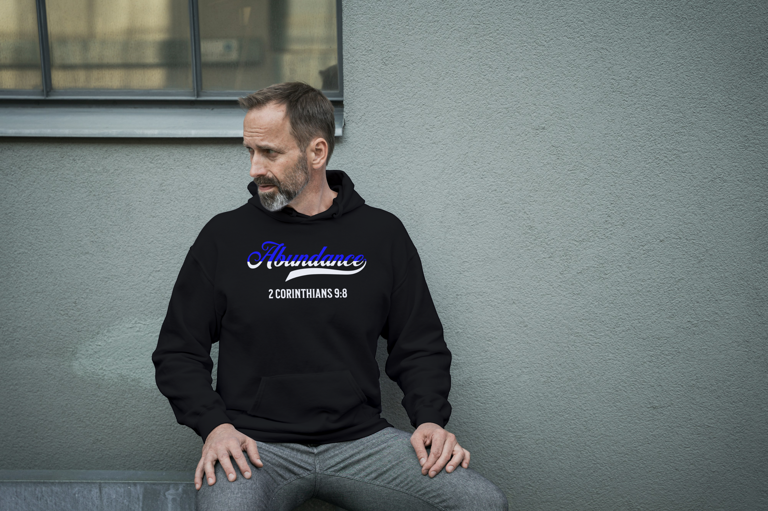 A man sitting against a gray wall wearing a black hoodie with the word 'Abundance' and the Bible verse '2 Corinthians 9:8' printed on it.