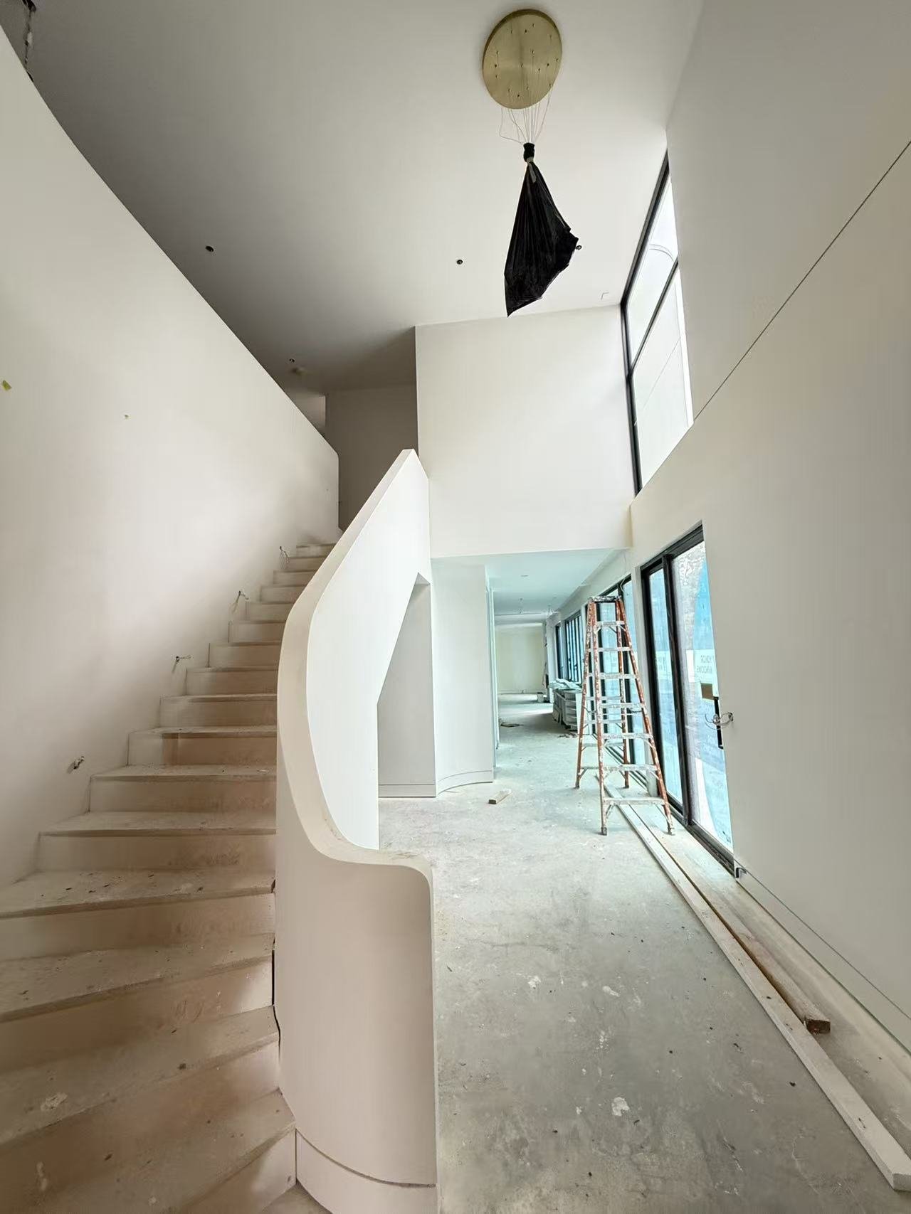 Interior of a modern, minimalist home under construction, featuring a curved staircase, large windows, and a ladder, with an unfinished floor.