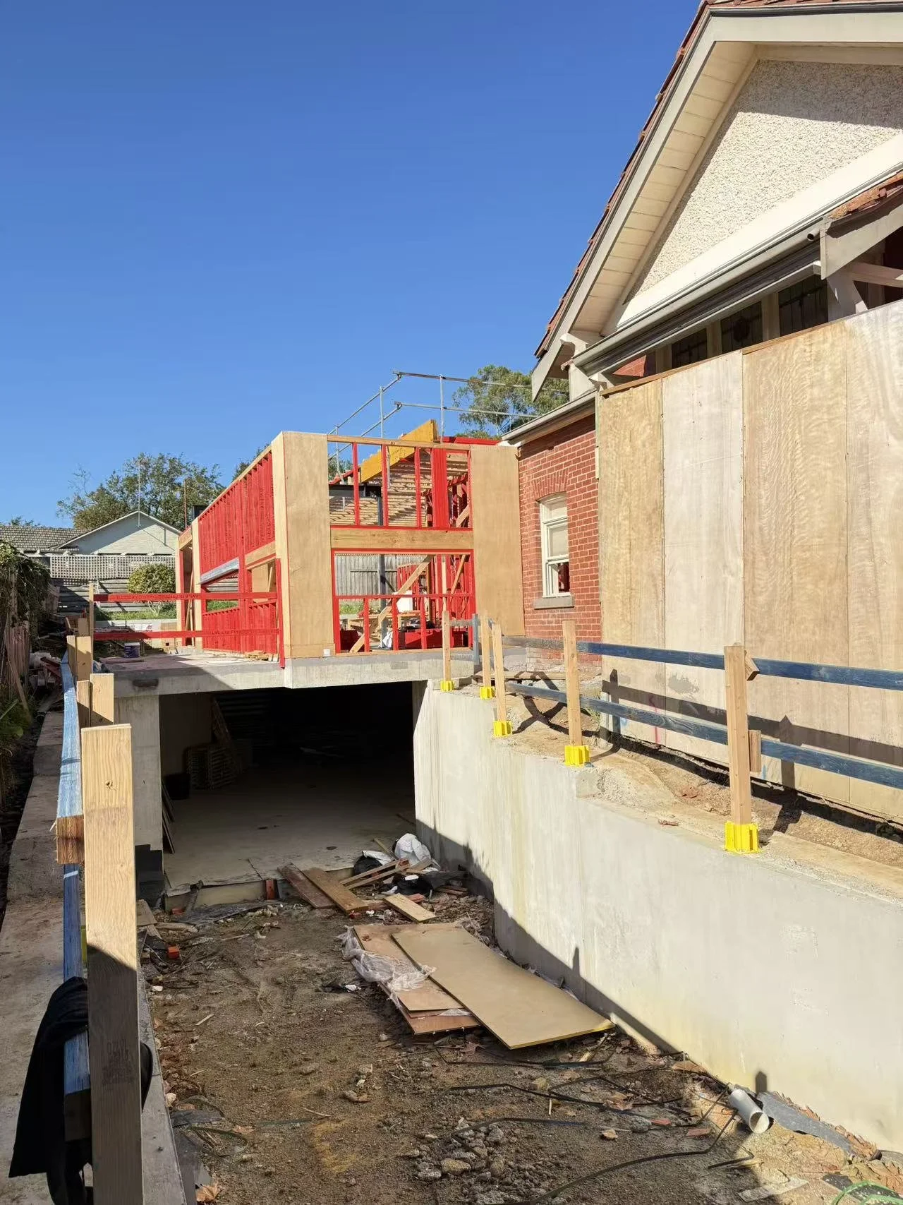 Construction site with a house under renovation, showing wooden framing, an unfinished deck with red railing, and a concrete garage. The ground is scattered with construction debris, and the sky is clear and blue.