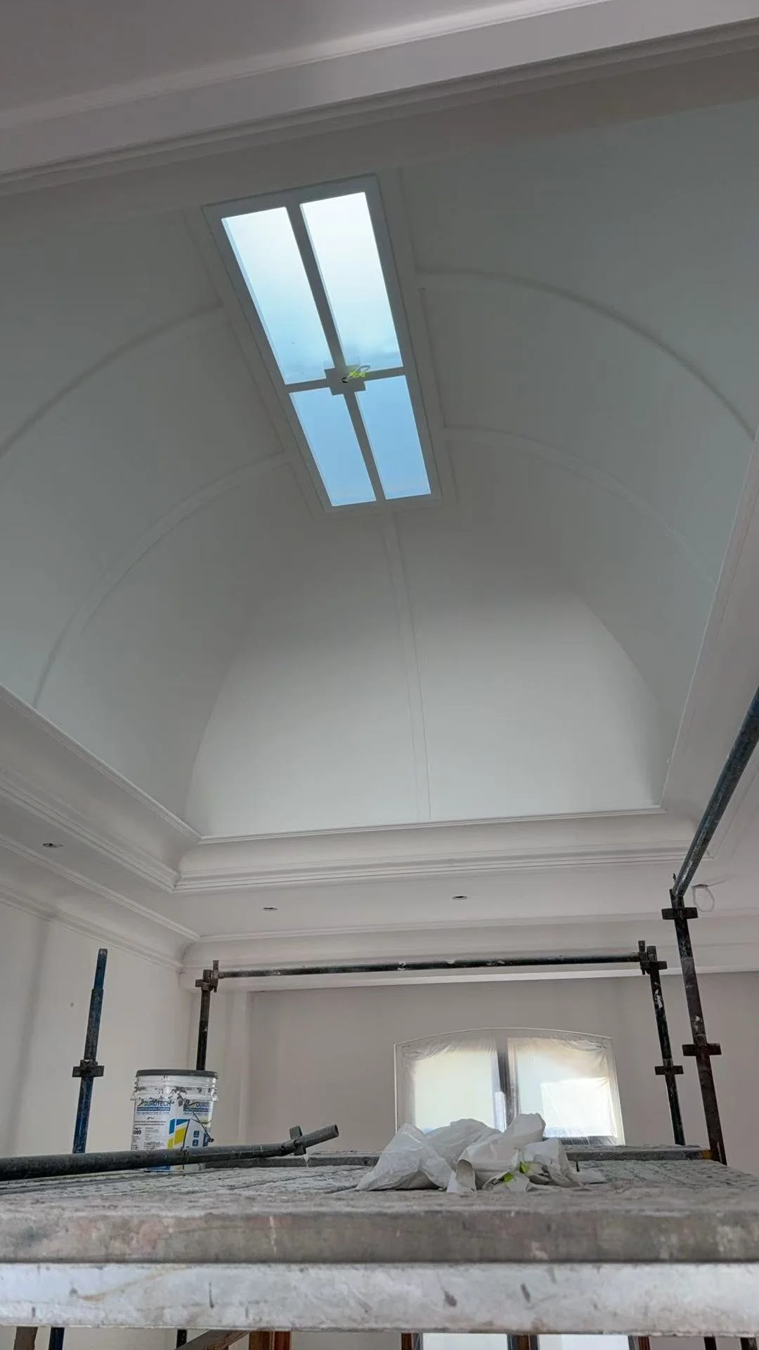 Interior room under renovation with scaffolding, a bucket, and painting supplies on a table, and an arched ceiling with a skylight.