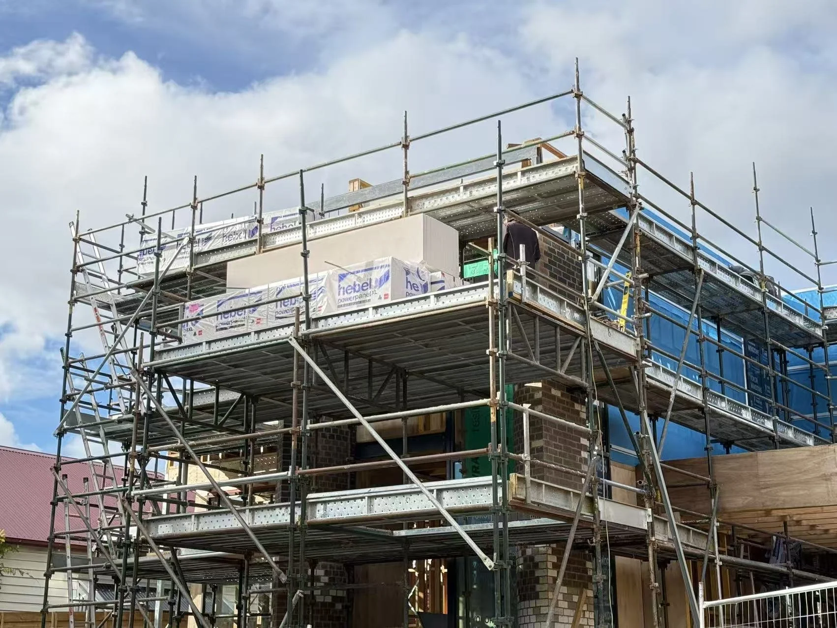 Building under construction with metal scaffolding and brick walls, partly covered with blue construction wrap, under a partly cloudy sky.