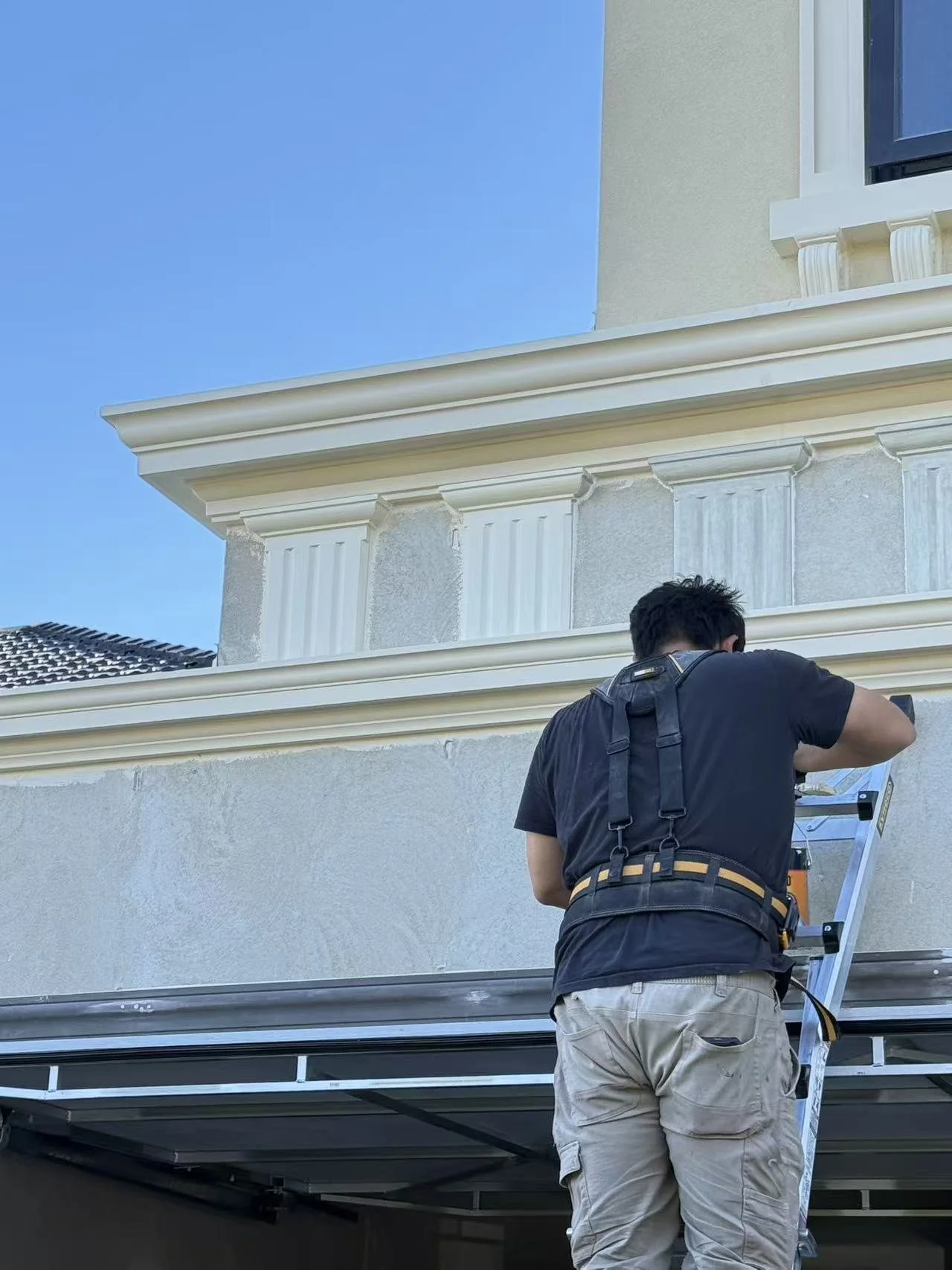 A person standing on a step ladder near a house, working on the exterior of the building on a sunny day.